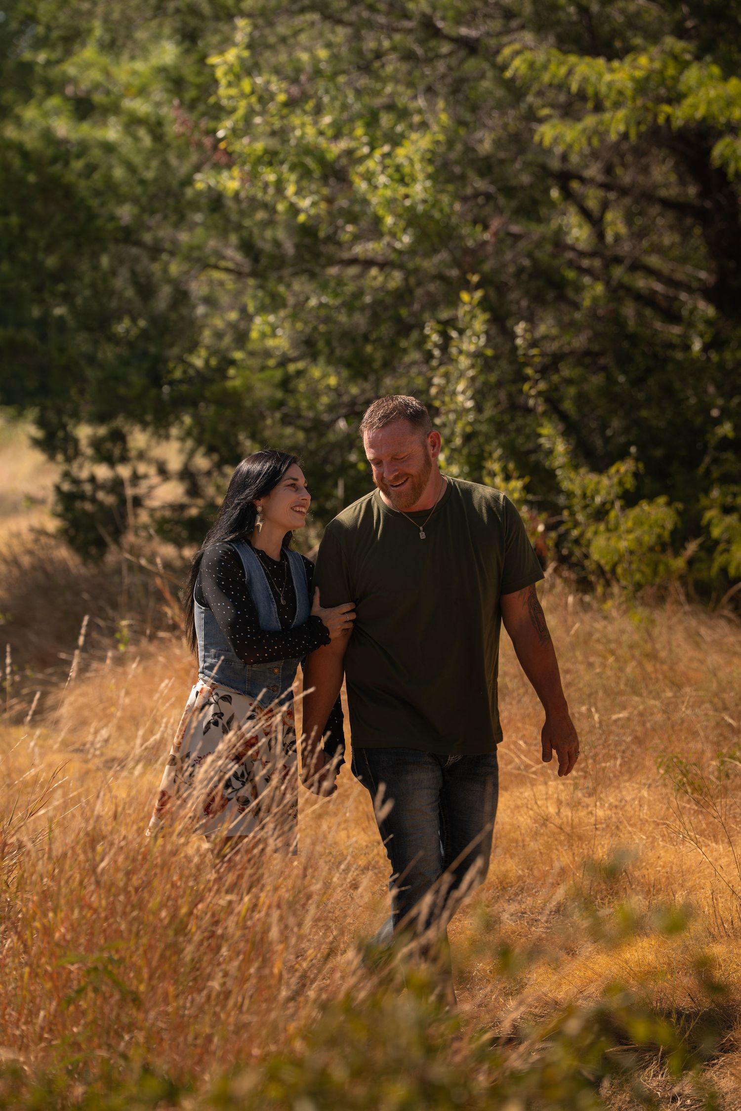 A couple walks hand-in-hand through golden meadow grass, smiling at each other with lush green trees in the background.