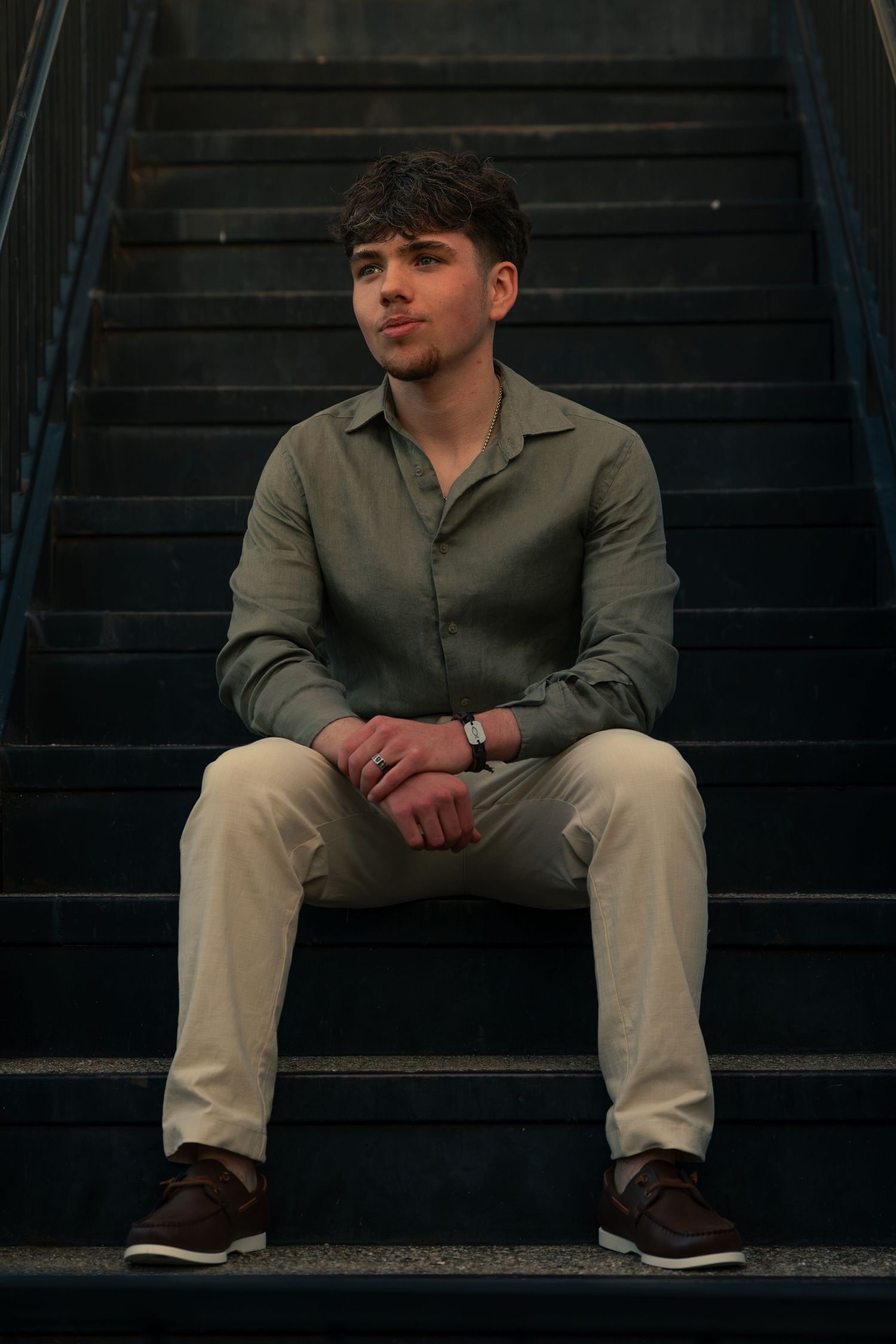 Young man in olive green shirt and khaki pants sitting on dark stairs, looking away.