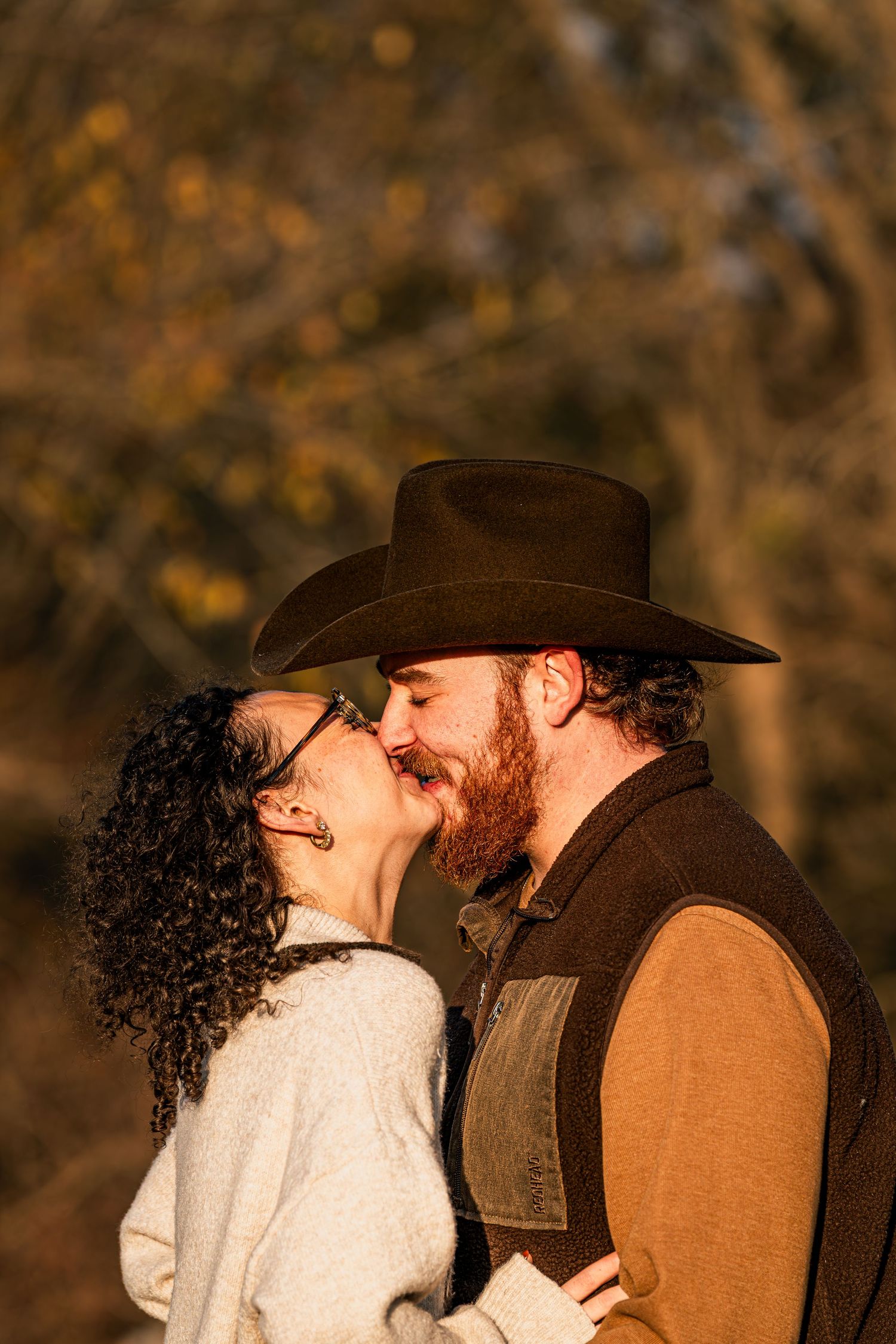 Bearded man in cowboy hat kissing curly-haired woman in warm autumn sunlight closeup.