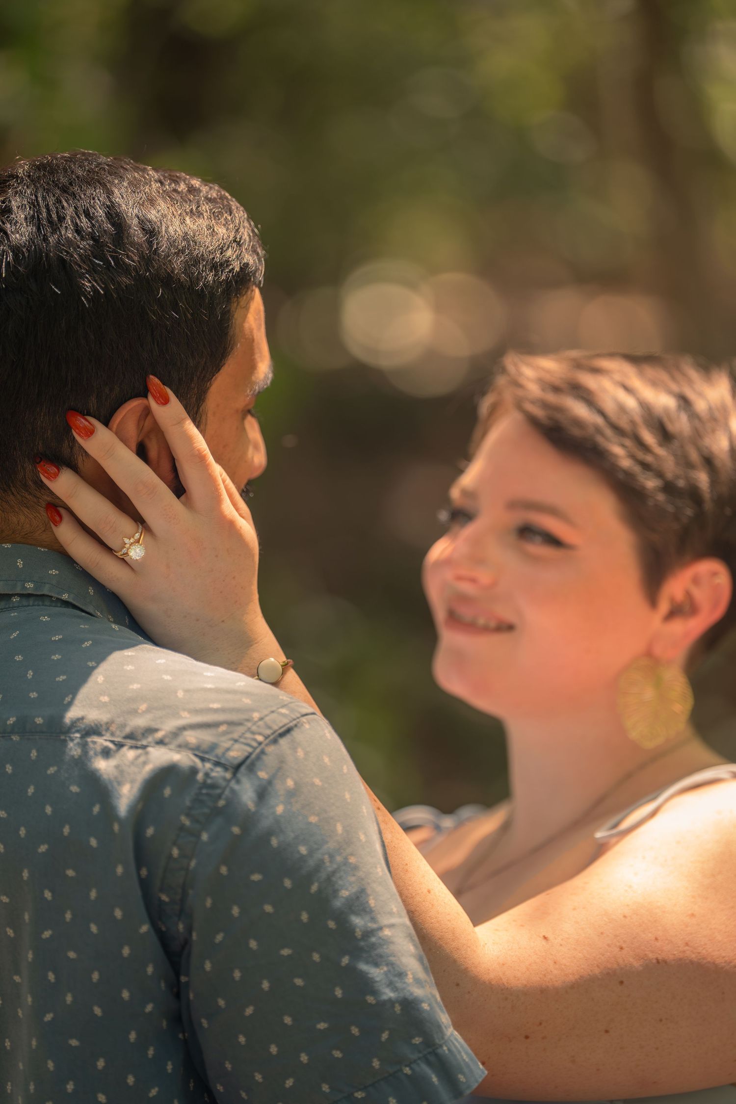 A smiling woman with short hair and gold earrings tenderly touches a mans face outdoors in warm sunlight.