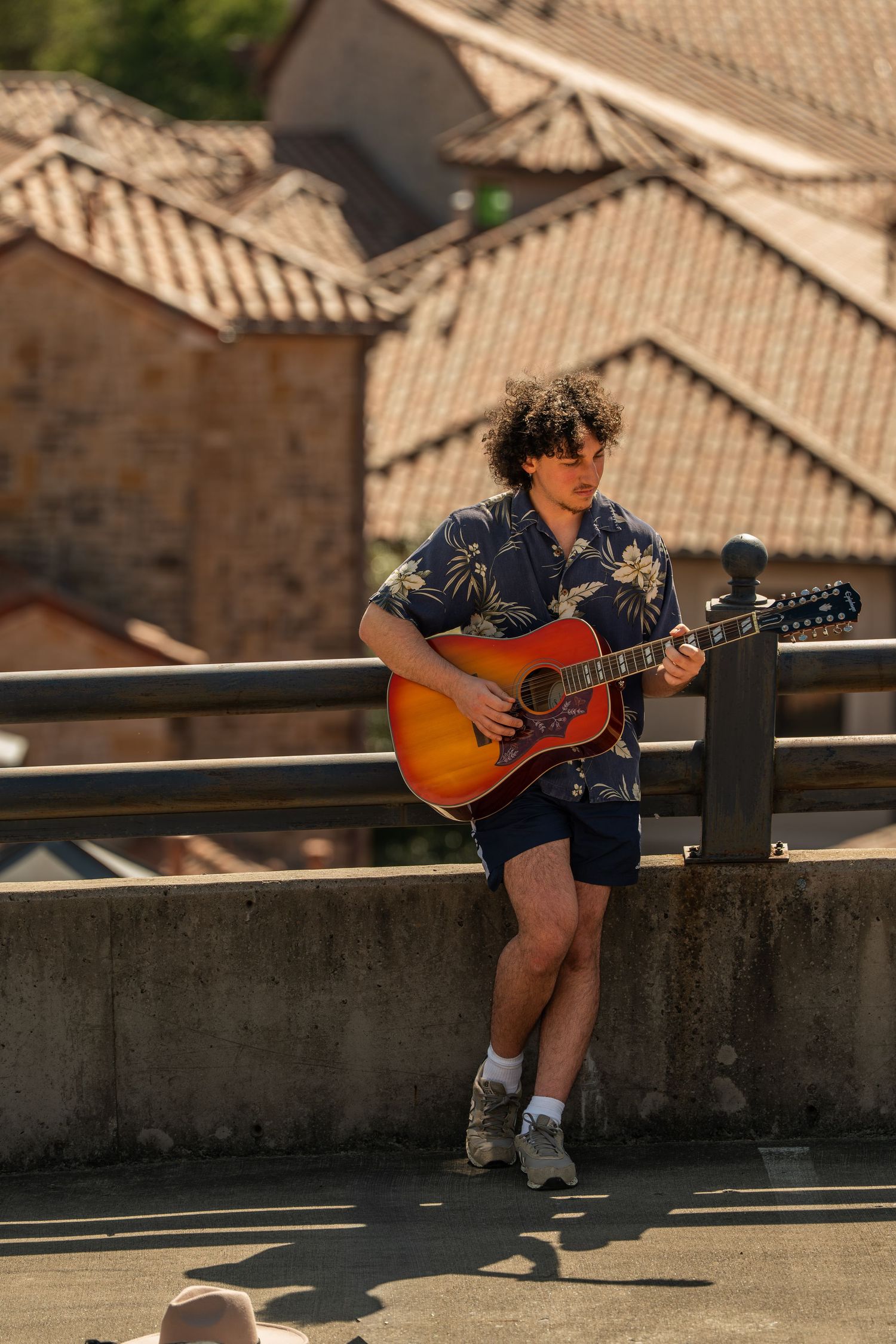 Young man playing acoustic guitar outdoors on a bridge with rooftops in the background.