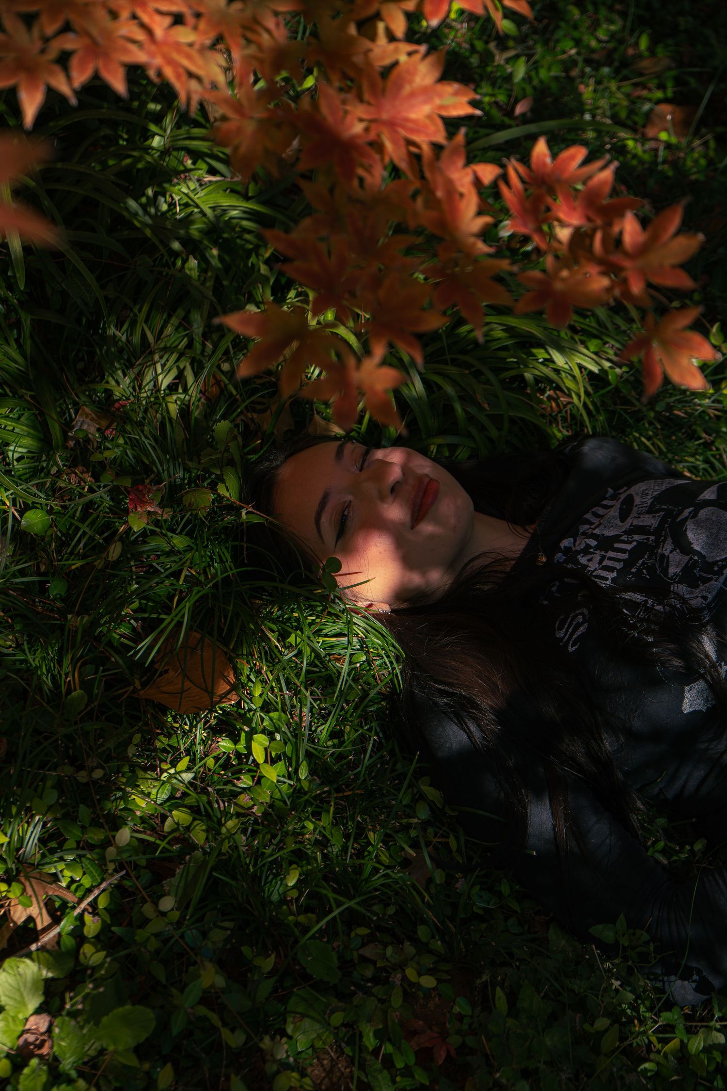 Young woman lying peacefully on green grass surrounded by orange autumn maple leaves in dark forest.