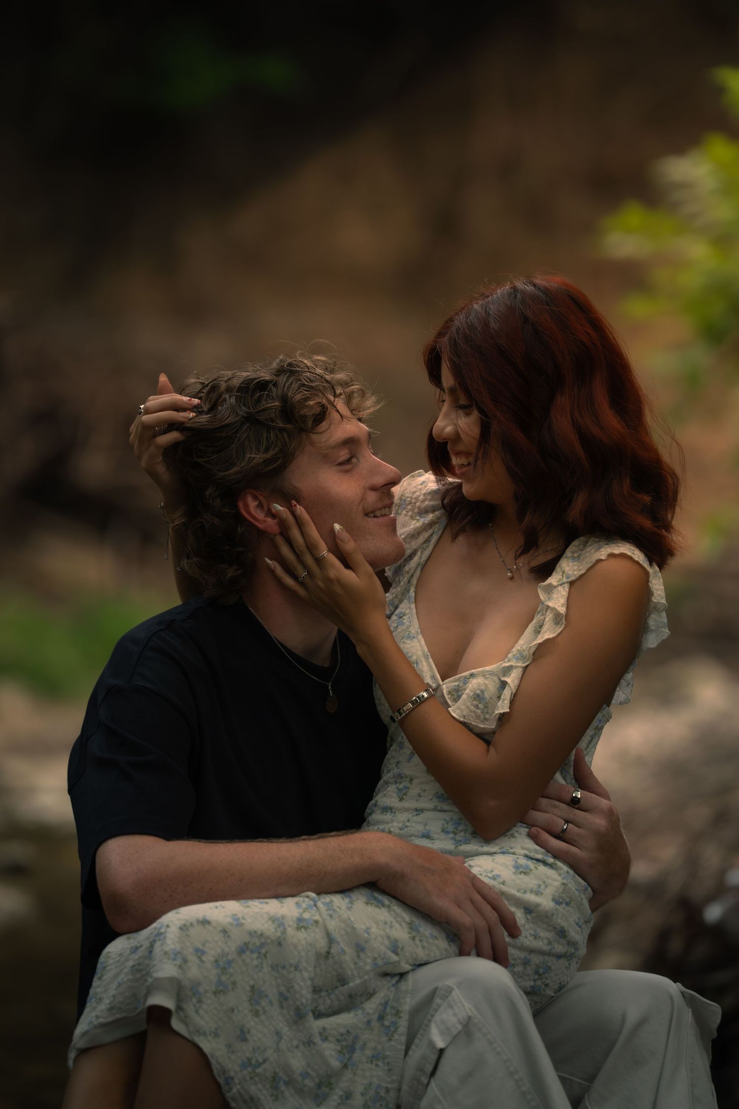 A couple shares a tender moment outdoors, woman in floral dress cradling man's face as they smile at each other.