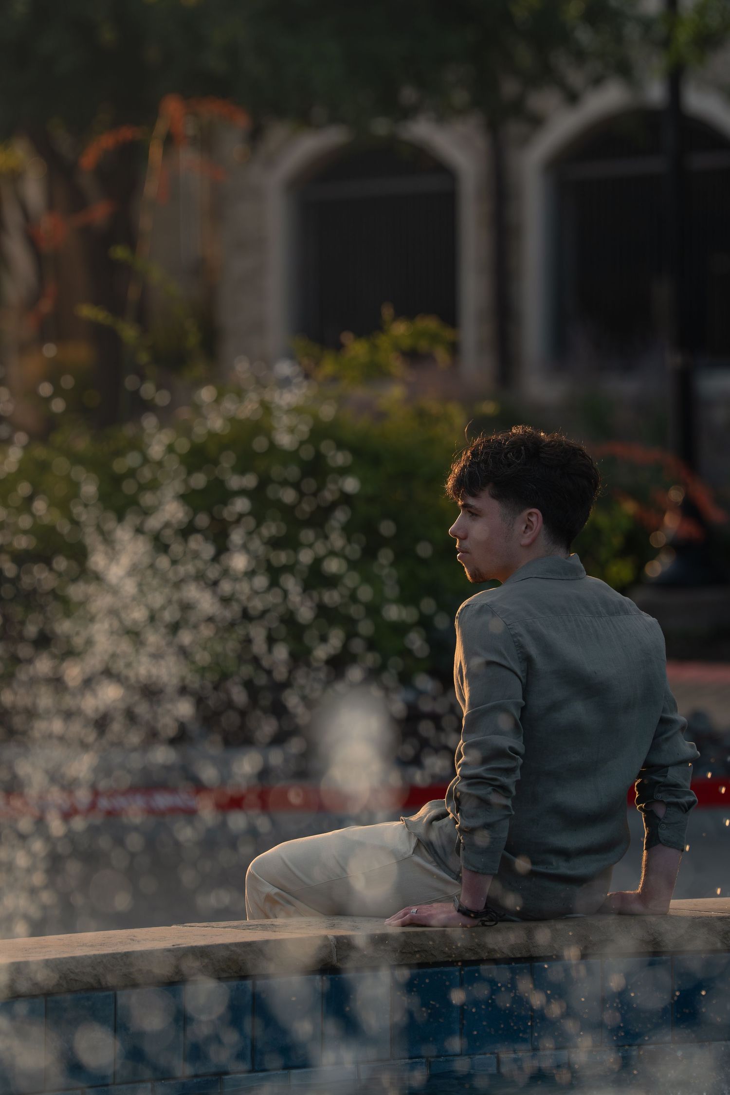Young man with curly hair sits pensively by a fountain in an urban plaza at golden hour.