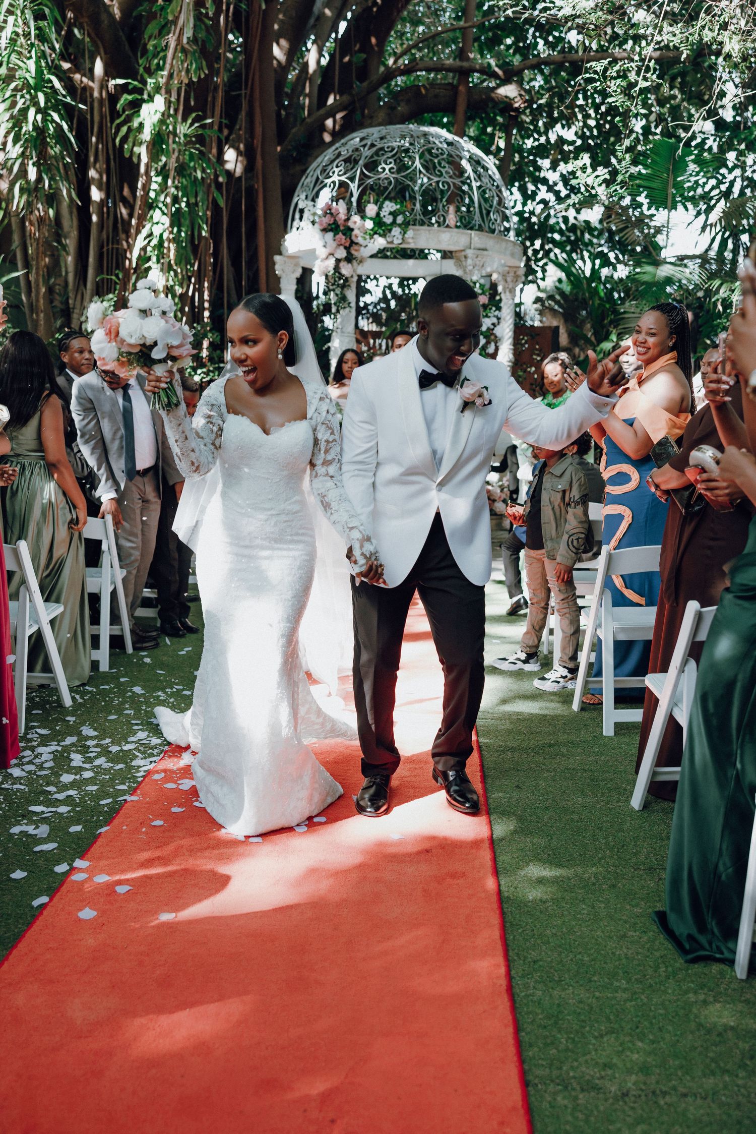 Bride and groom walk down red carpet aisle outdoors at garden wedding ceremony with guests cheering.