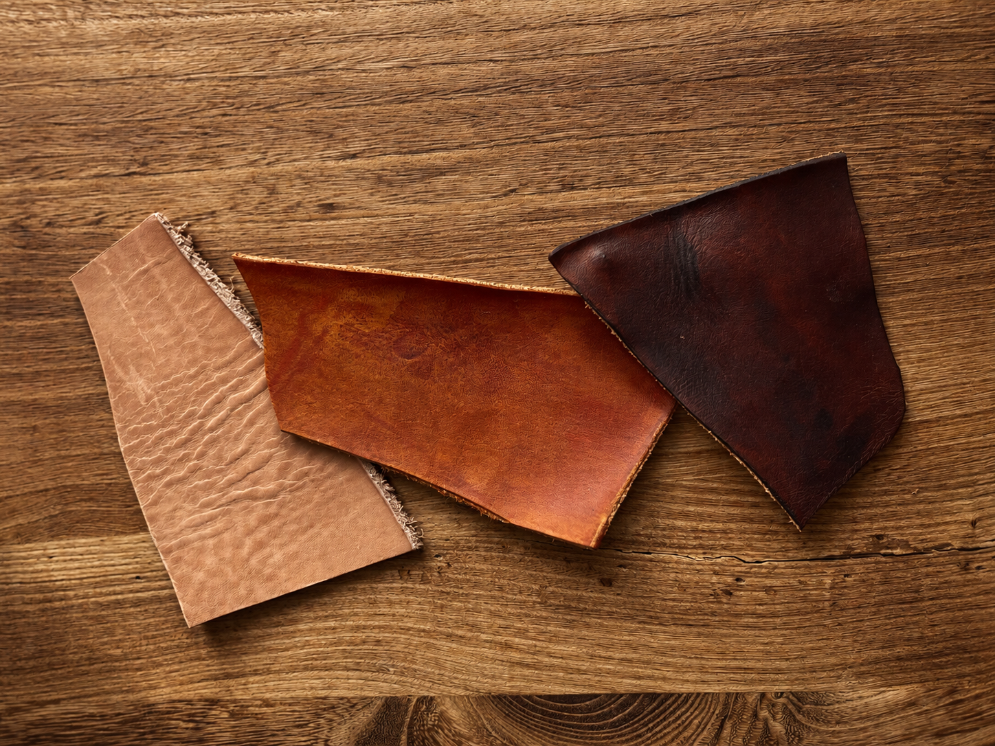 Flat lay of three leather pieces in tan, cognac, and dark brown tones arranged on a wooden background.