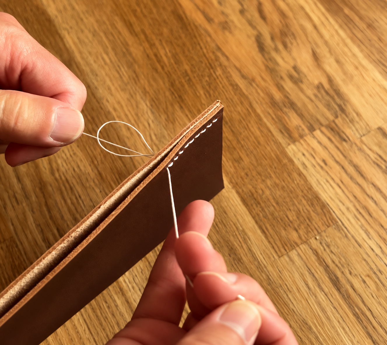 Hands stitching brown leather with white thread on a wooden surface, demonstrating traditional leather bookbinding craft technique.