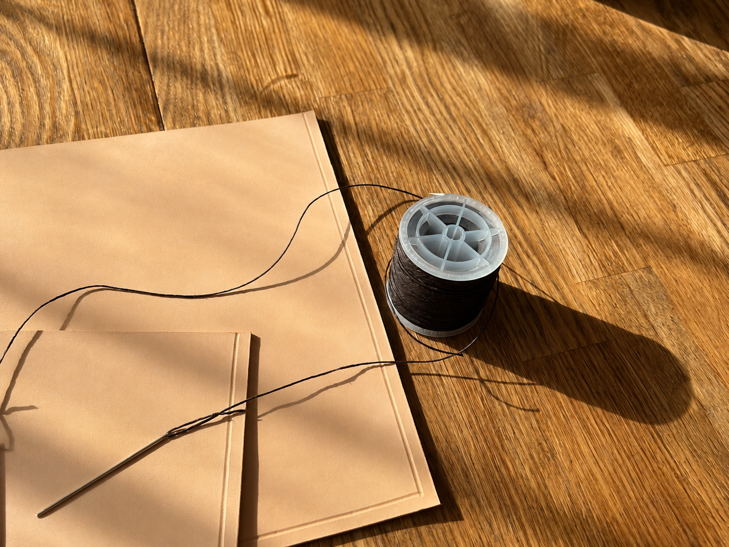 Black thread spool and needle on tan leather piece, bathed in warm sunlight on a wooden surface.