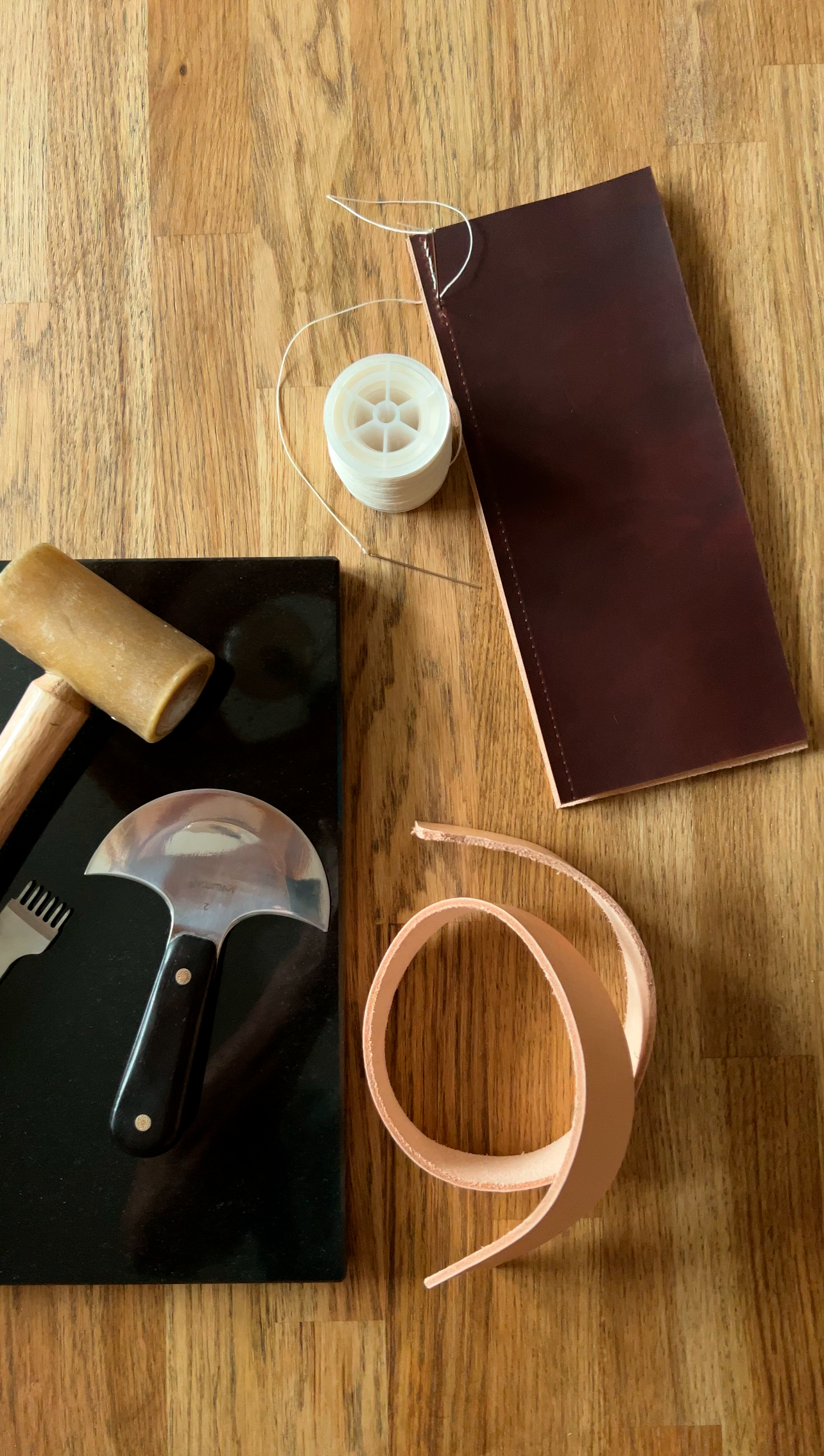 Leather crafting supplies including a mallet, thread, burgundy leather piece, and tan leather strip on wood.