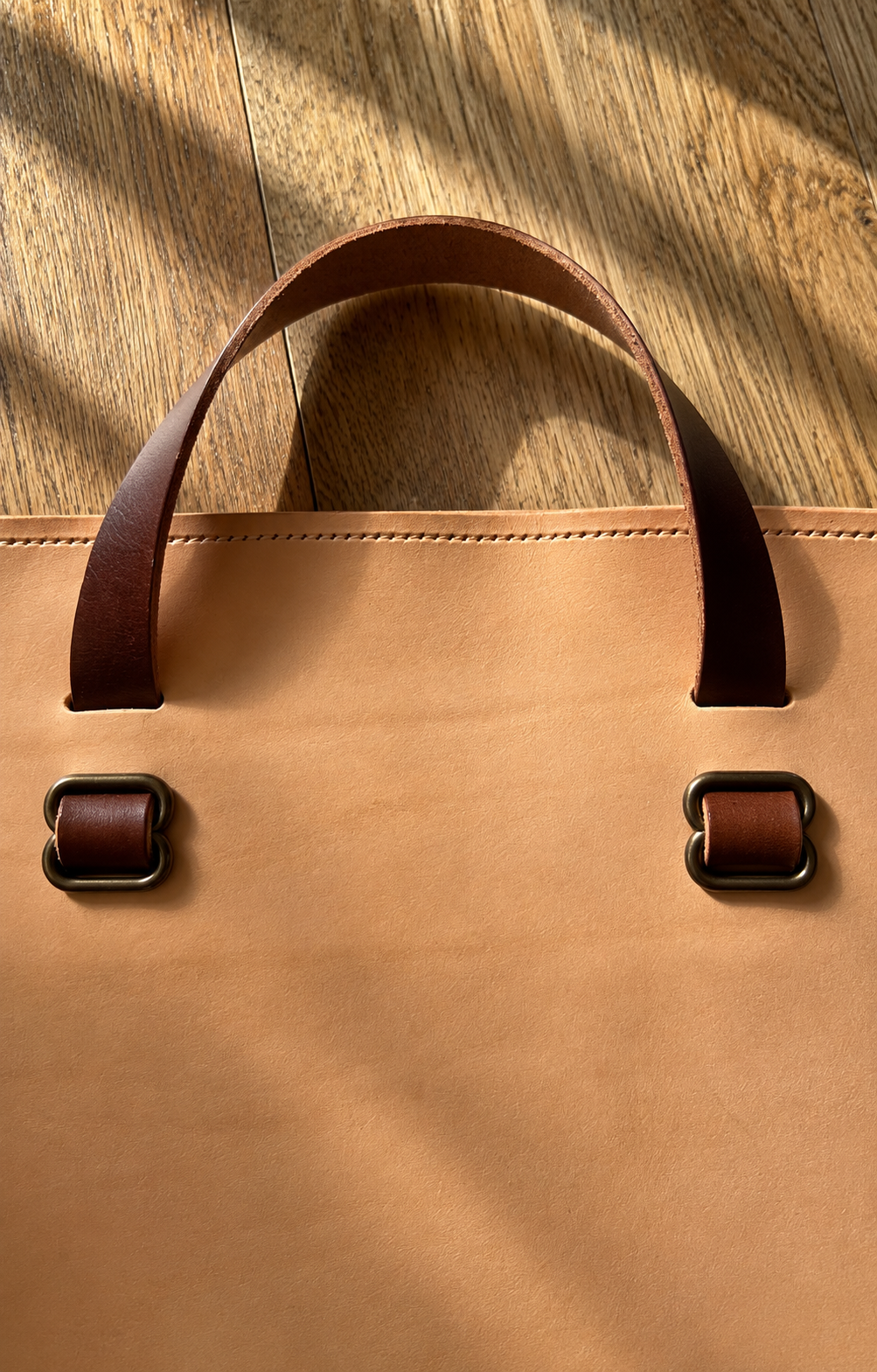 Close-up of a tan leather tote bag with dark brown handles and silver buckle hardware on a wooden surface.