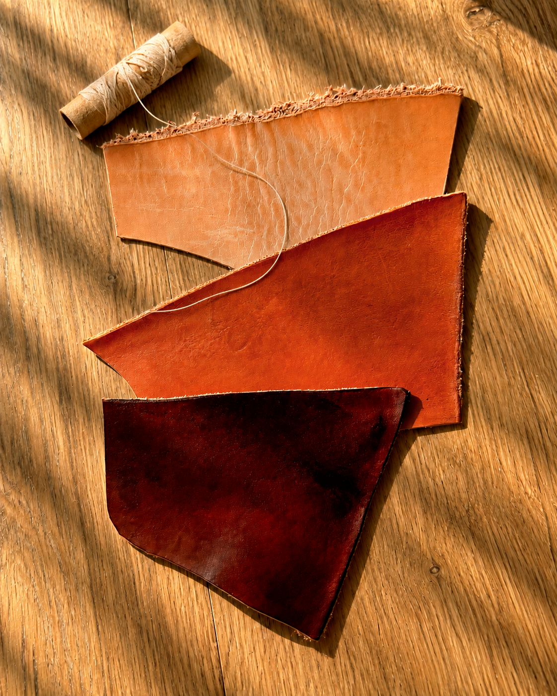 Three leather swatches in tan, reddish-brown, and dark burgundy arranged on a wooden surface with a spool of twine.