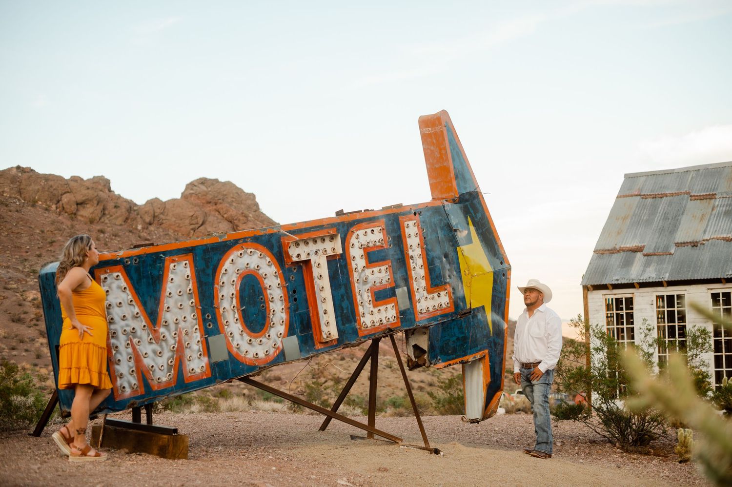 Engaged couple stands beside a vintage neon Motel sign in a desert landscape at dusk.