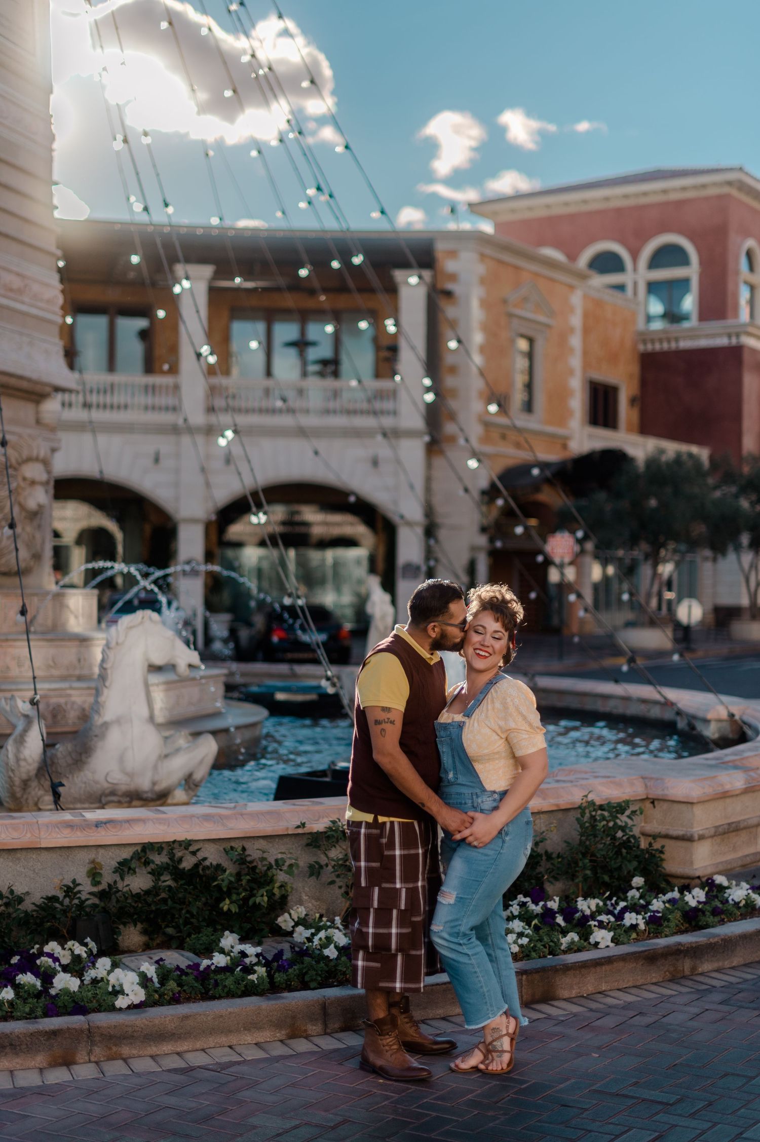 Couple posing by a fountain with string lights at an Italian-themed Las Vegas plaza in daylight.