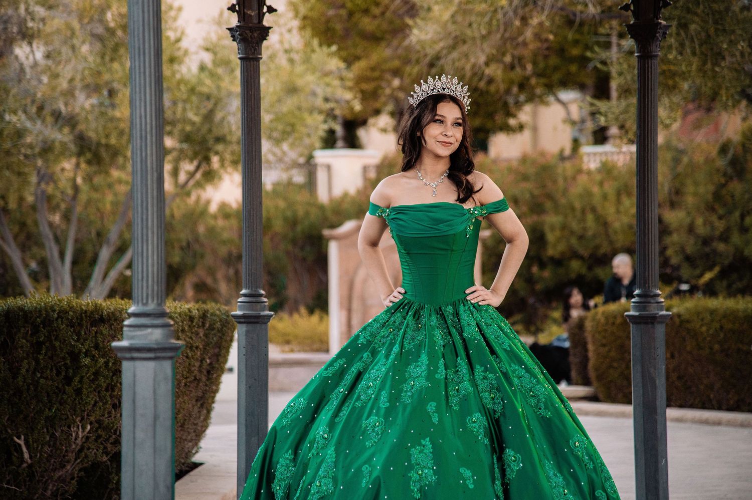 Teen girl in green off-shoulder quinceañera ball gown and tiara posing between outdoor pillars.
