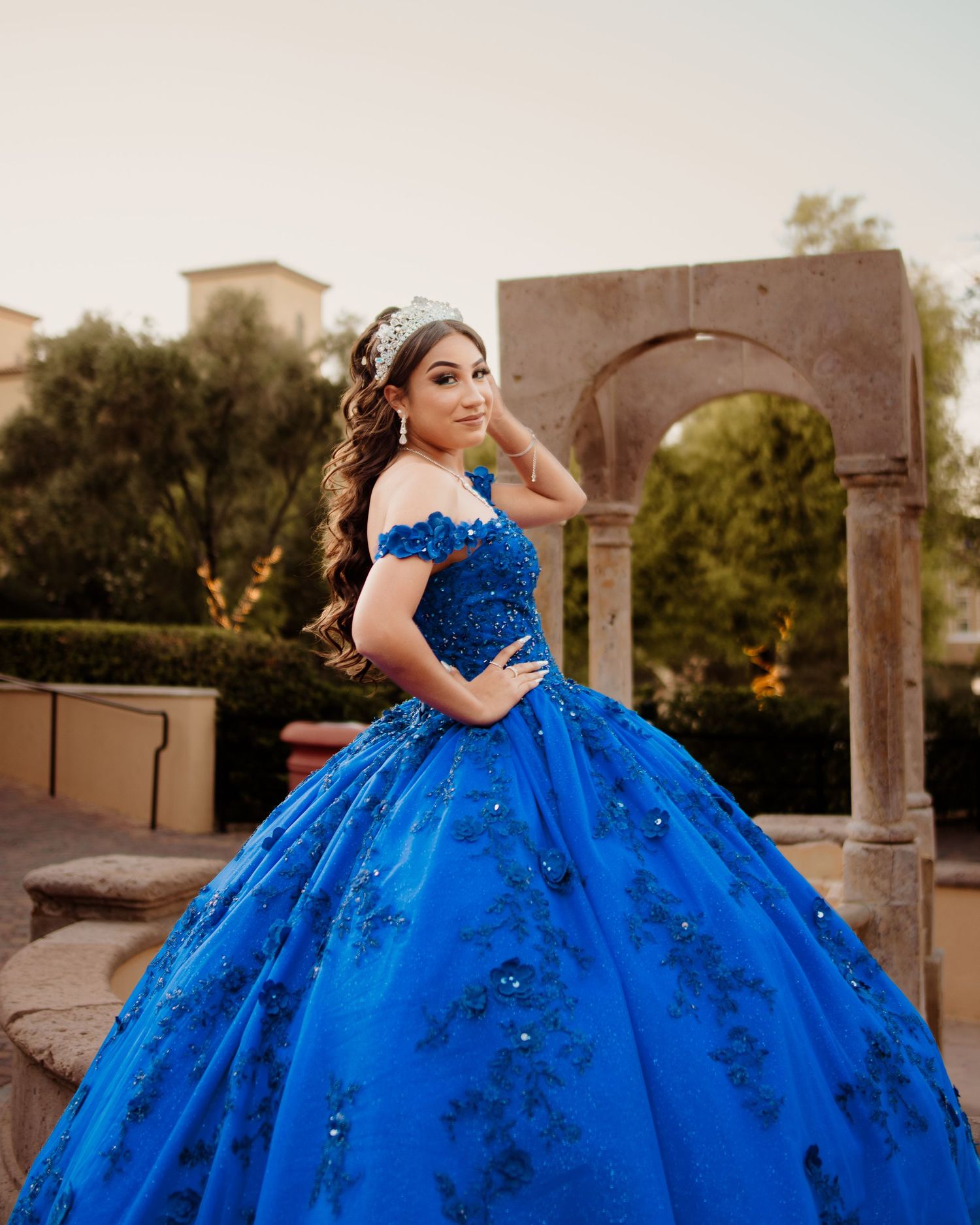 Young woman in royal blue off-shoulder quinceañera gown with crystal tiara poses at golden hour outdoors.