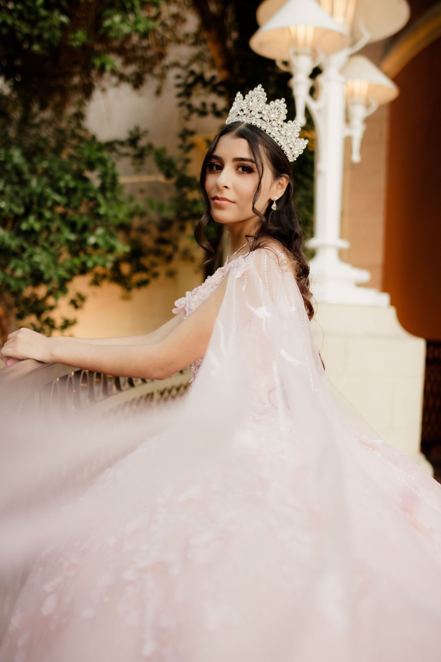 Young woman in blush pink quinceañera ball gown with floral cape and crystal crown poses elegantly indoors.