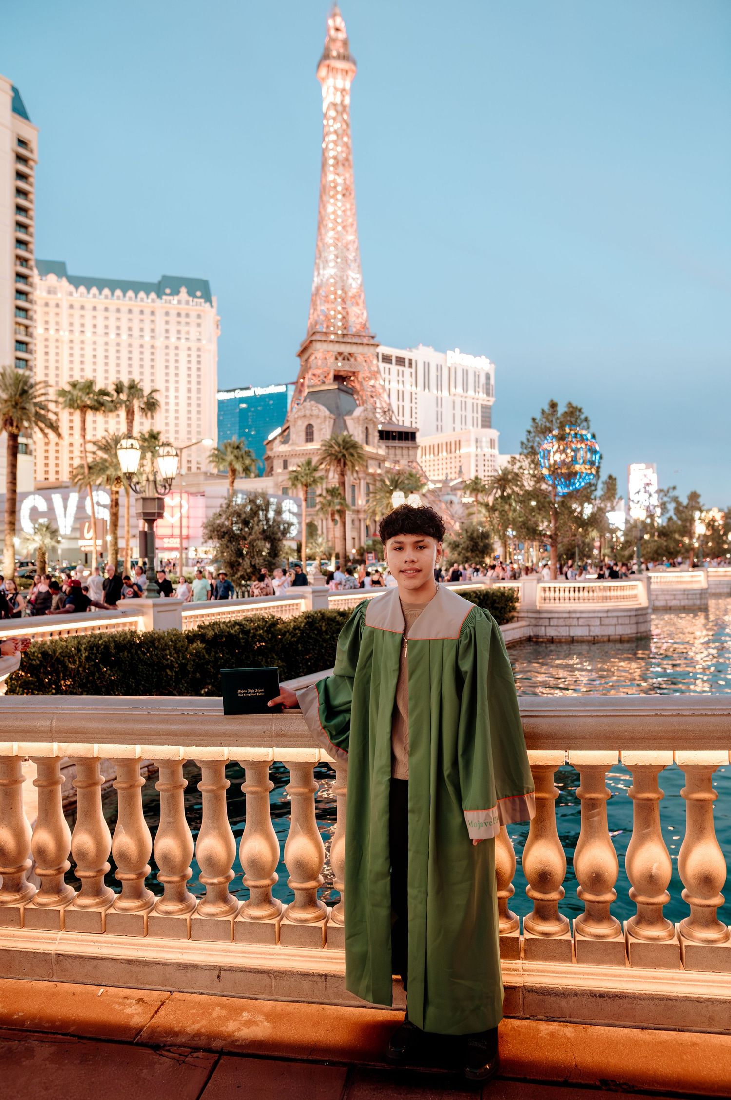 Smiling graduate in green gown stands before Paris Las Vegas Eiffel Tower replica at twilight.