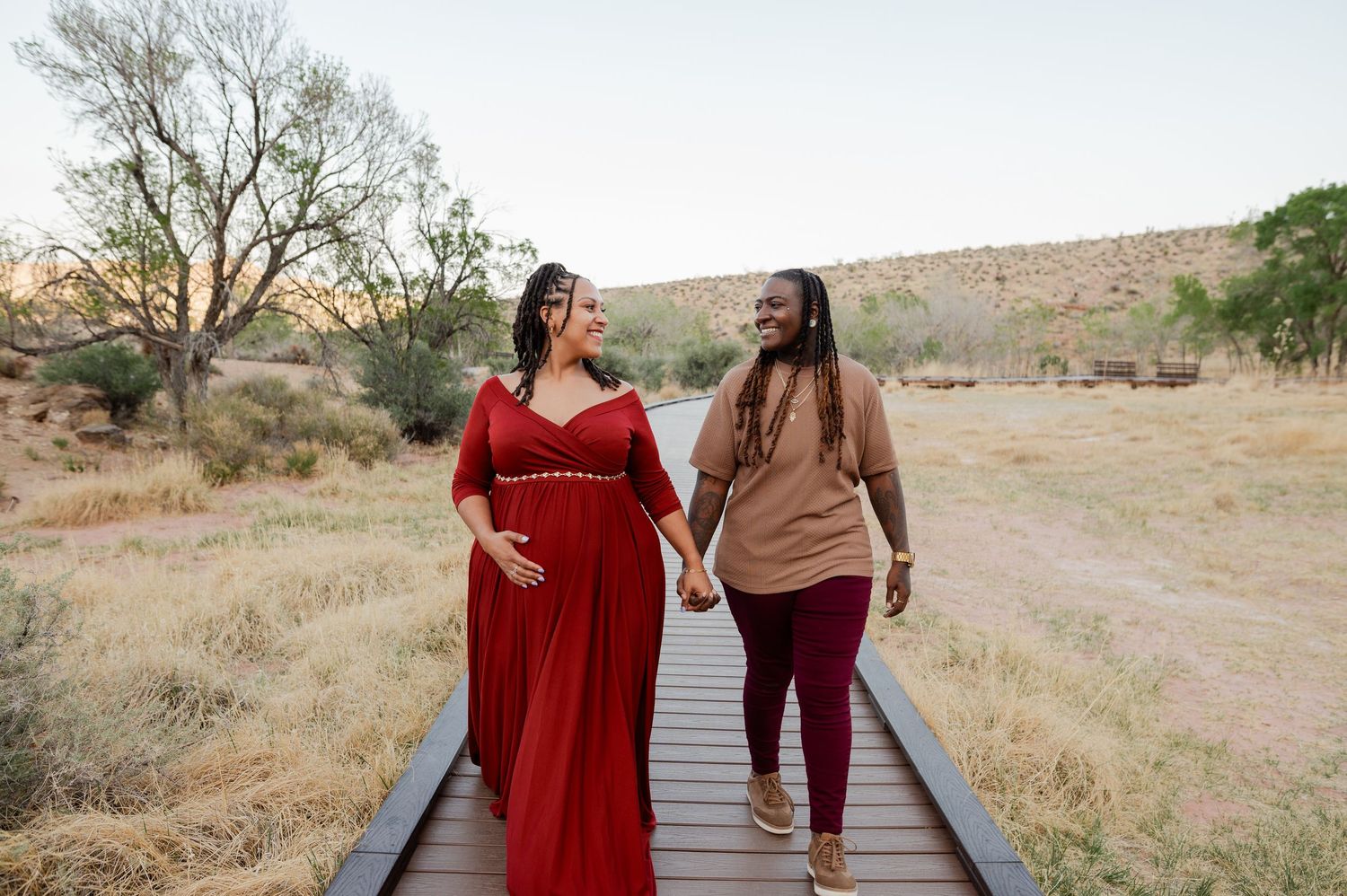 Pregnant woman in red gown and partner walk hand-in-hand on wooden boardwalk in nature.