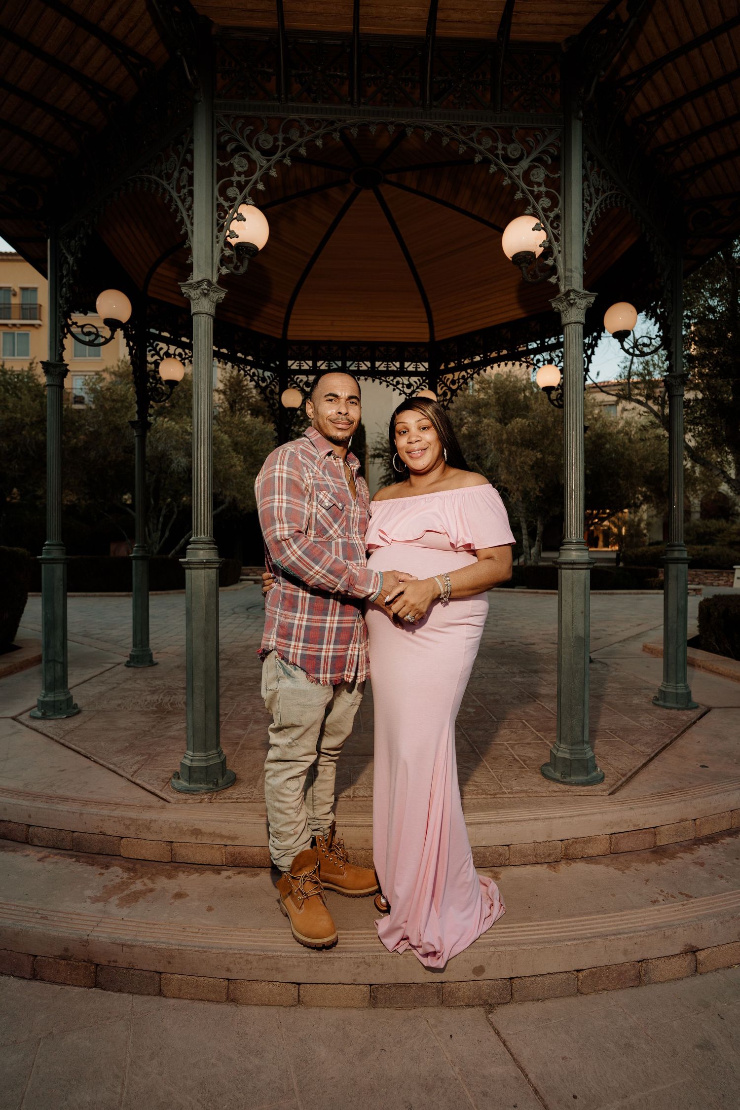 Couple posing under ornate gazebo at night, pregnant woman in pink maternity gown held by her partner.