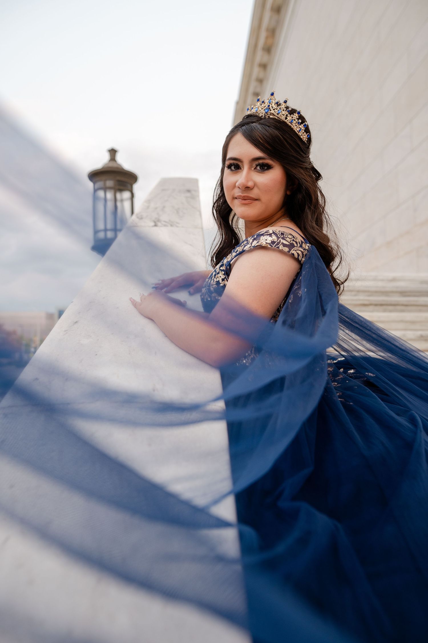 Young woman in a navy blue quinceañera gown with a flowing skirt and tiara poses elegantly outdoors.