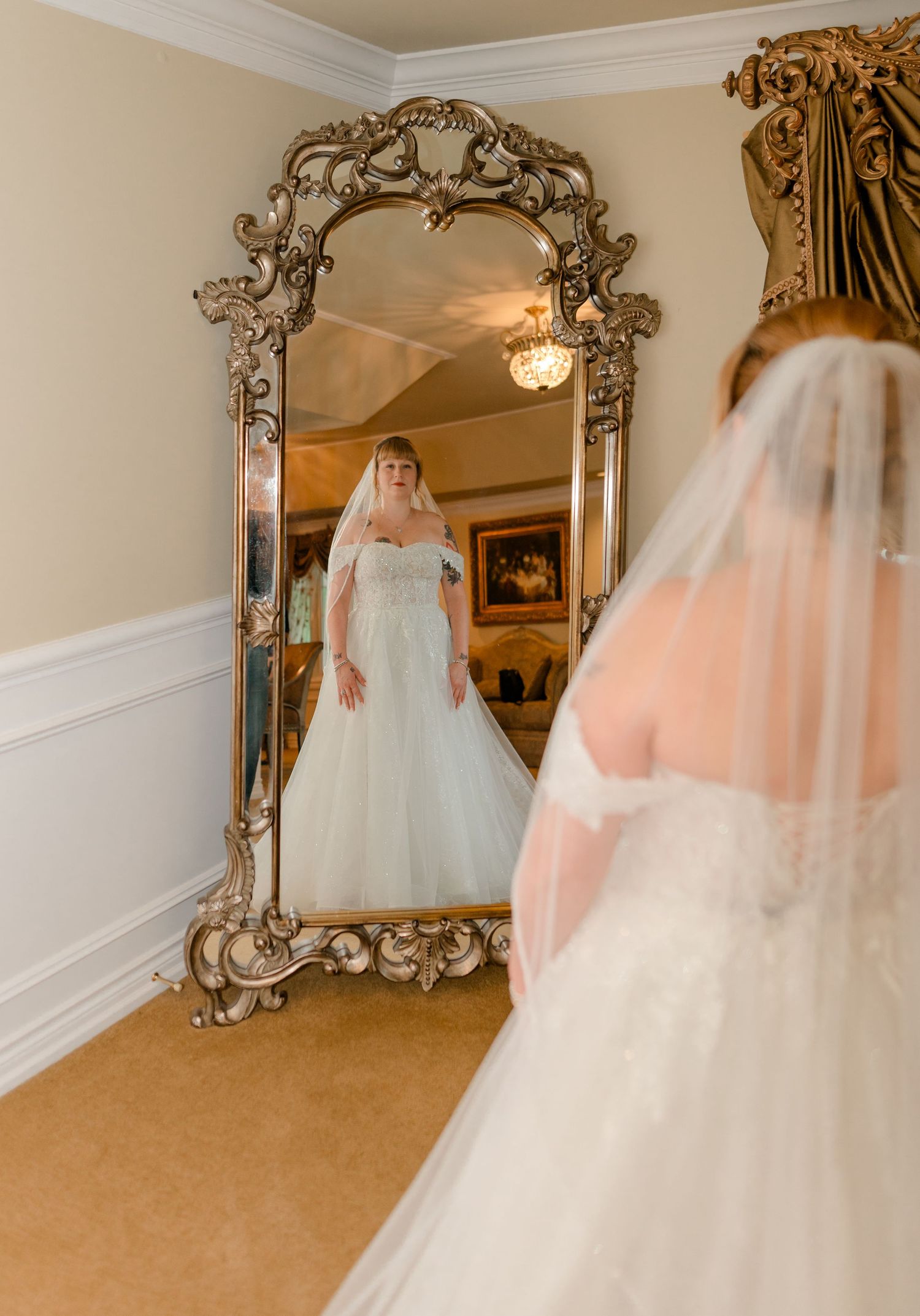 Bride in a white ball gown and veil admires her reflection in an ornate gold floor mirror.
