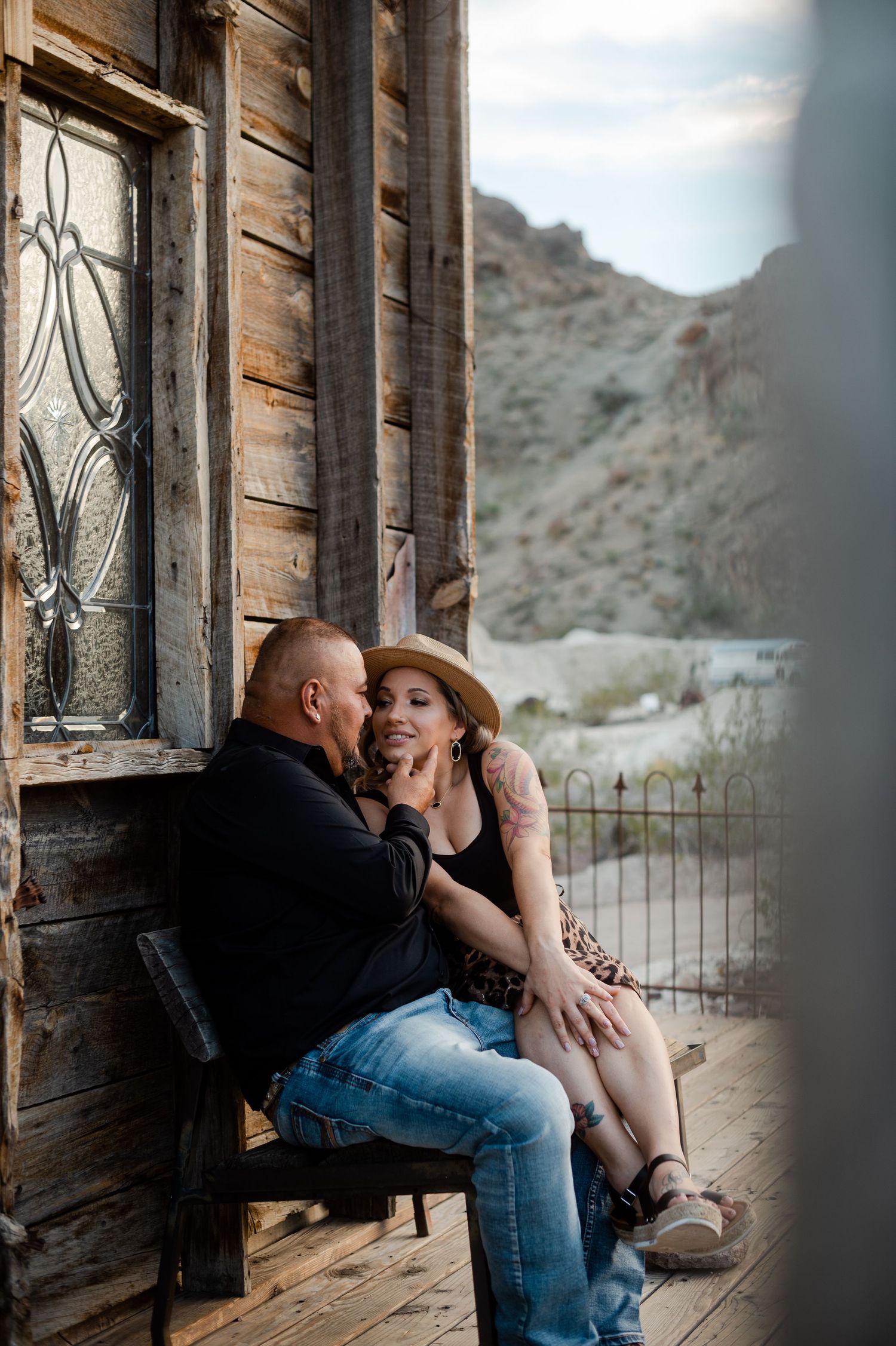 Couple sitting close together on a weathered wooden porch, smiling intimately near an ornate window.