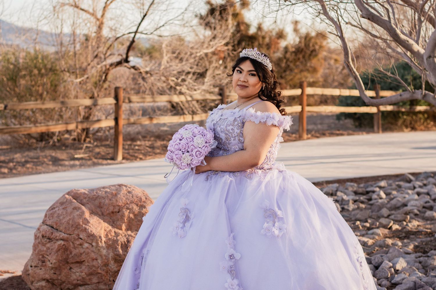 Young woman in lavender quinceañera ball gown with floral appliques holds bouquet outdoors.