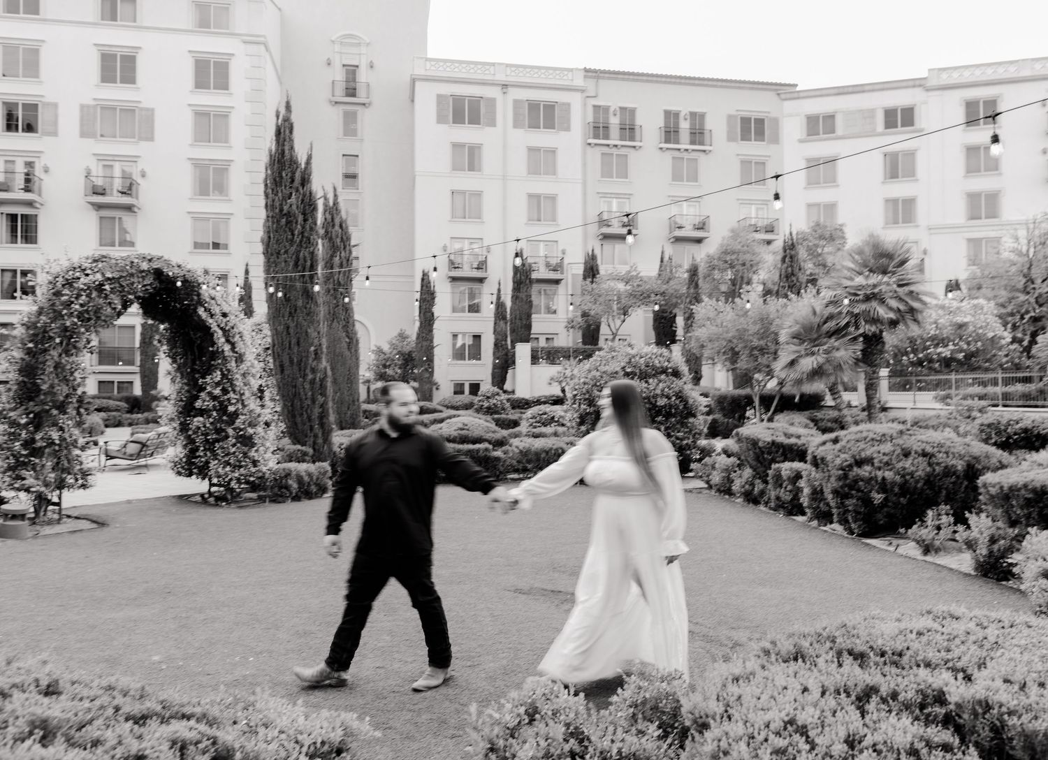 Black and white photo of bride and groom holding hands in a lush garden courtyard.
