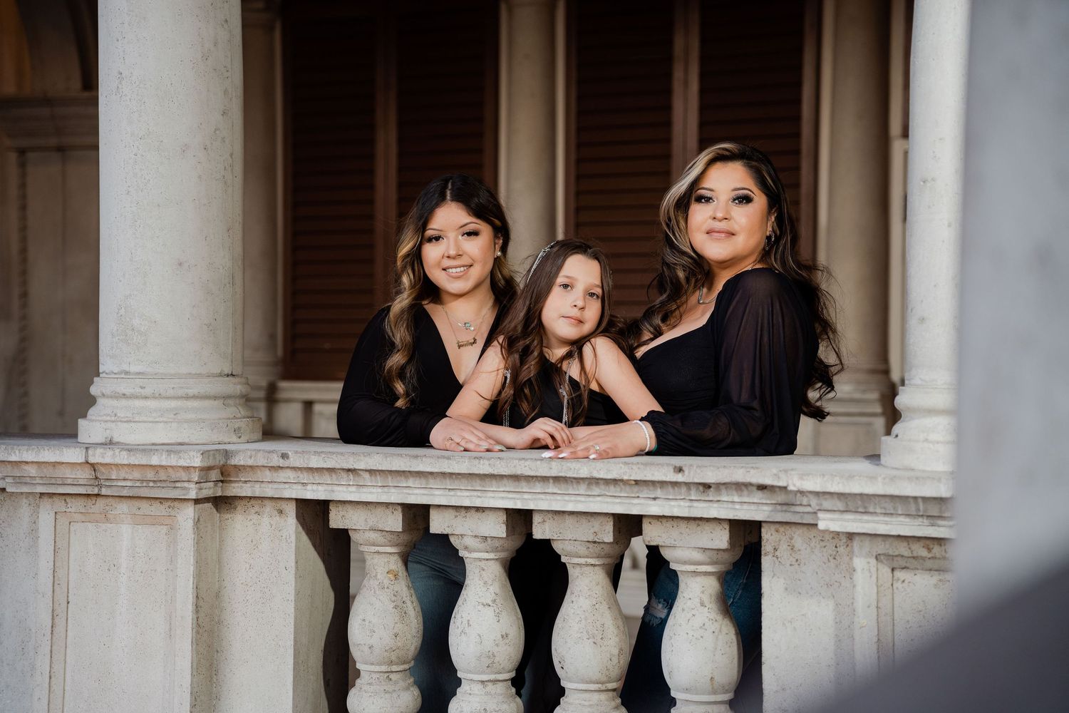 Three women in black outfits pose together on an elegant stone balcony with classical columns.