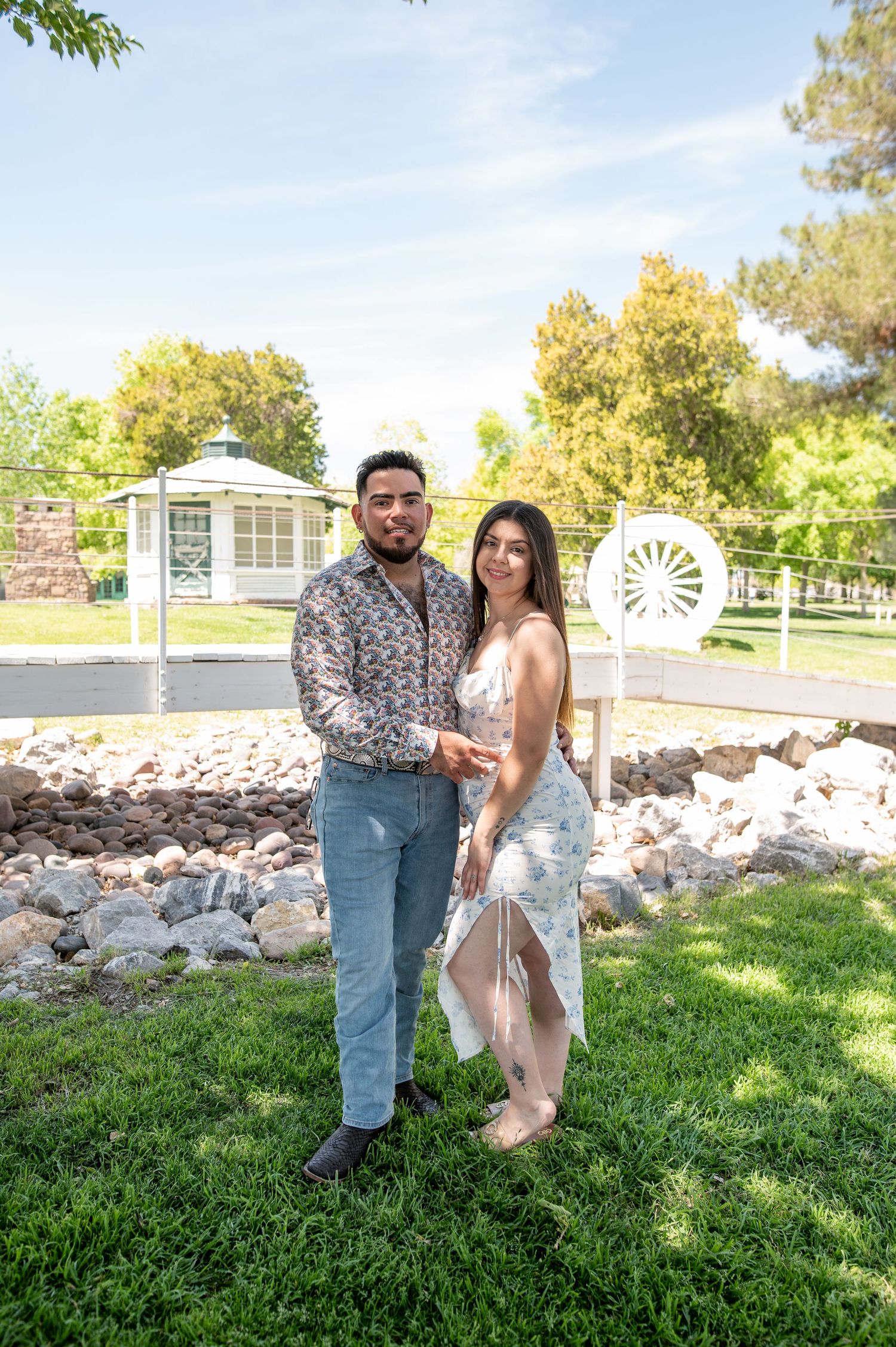 A couple poses together in a lush green park with a gazebo and decorative wagon wheel in the background.