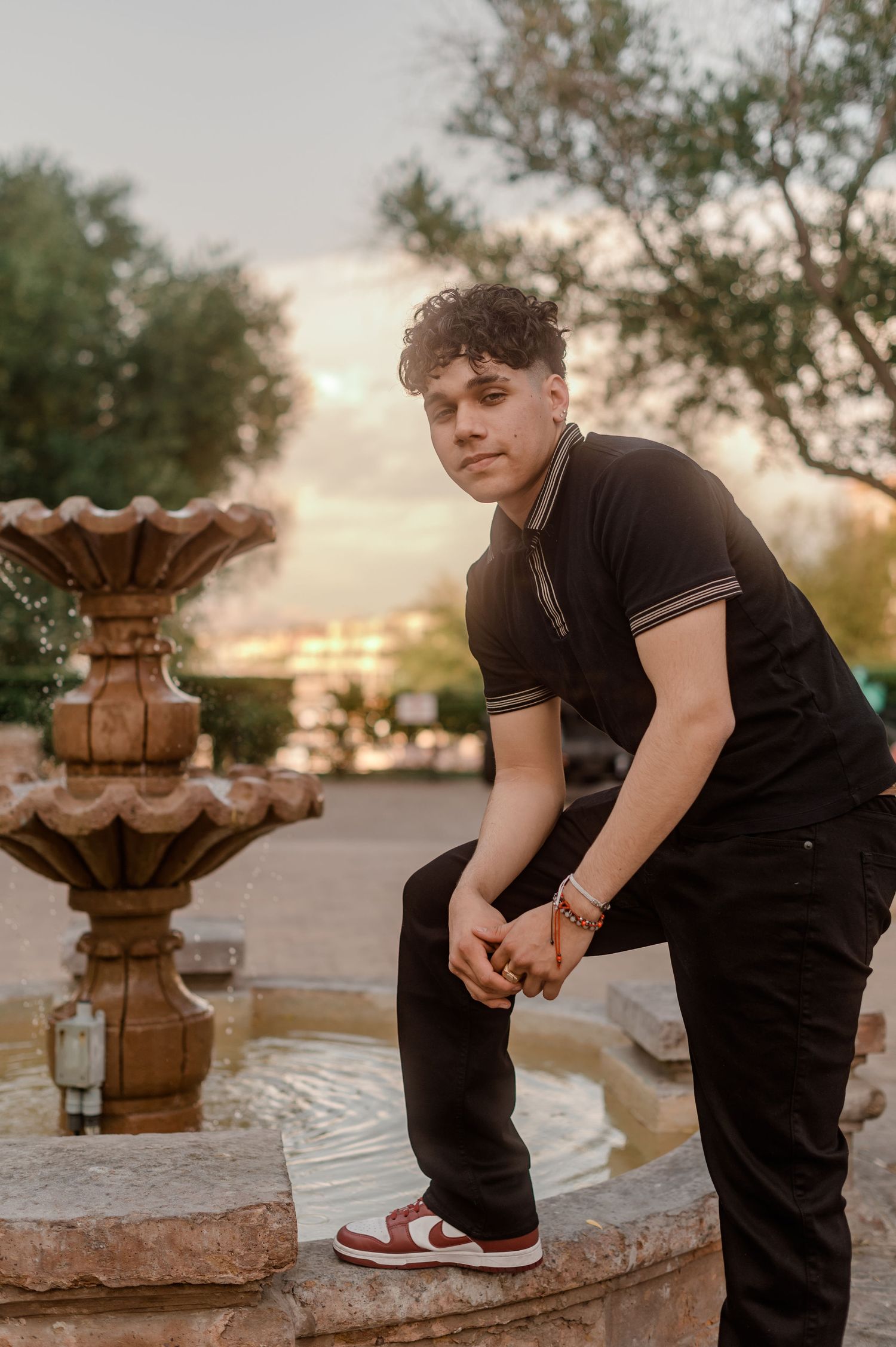 Young man in black polo sits on stone fountain edge at sunset in garden setting.