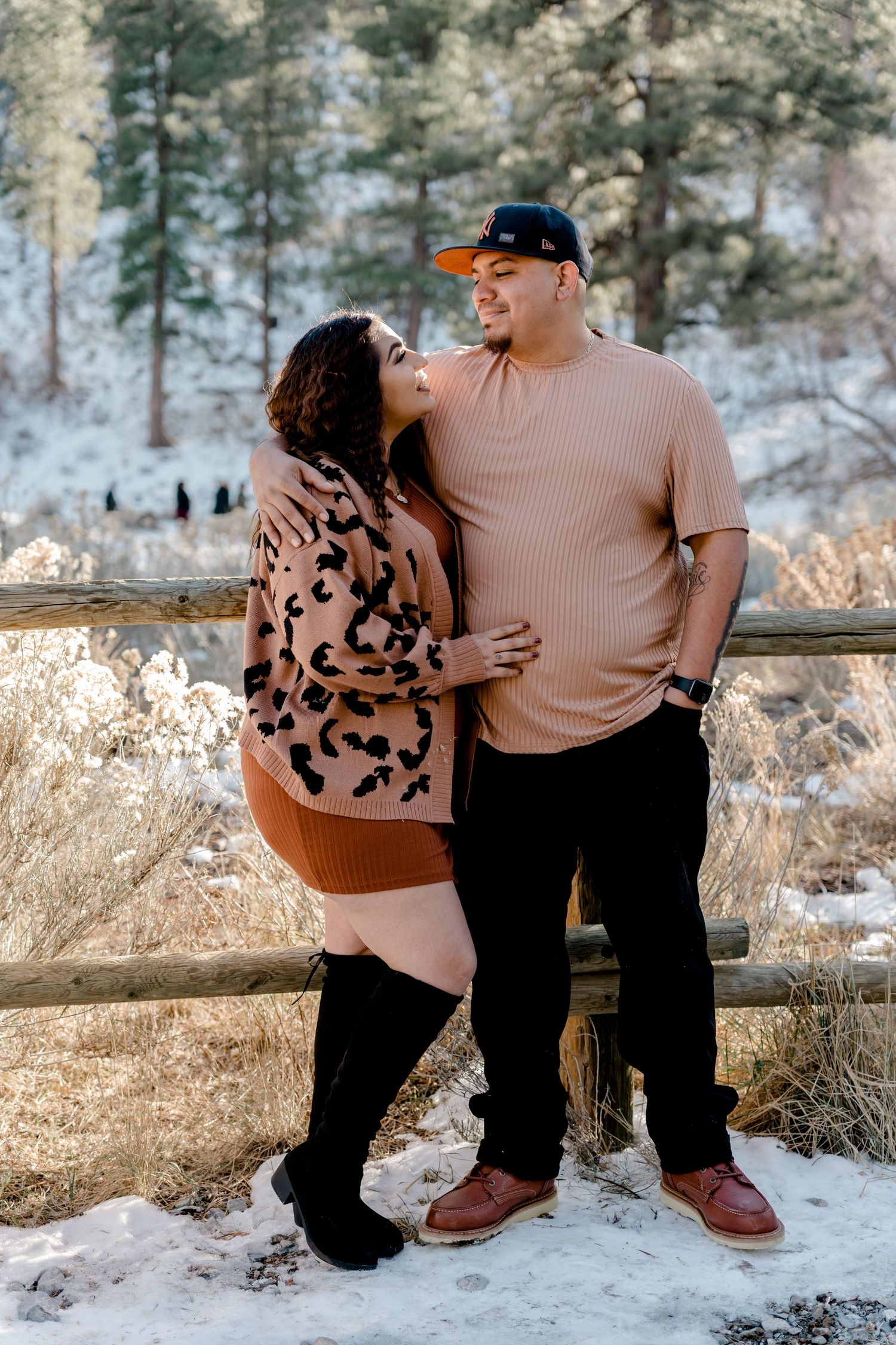 Couple embracing near rustic wooden fence in snowy mountain forest setting.
