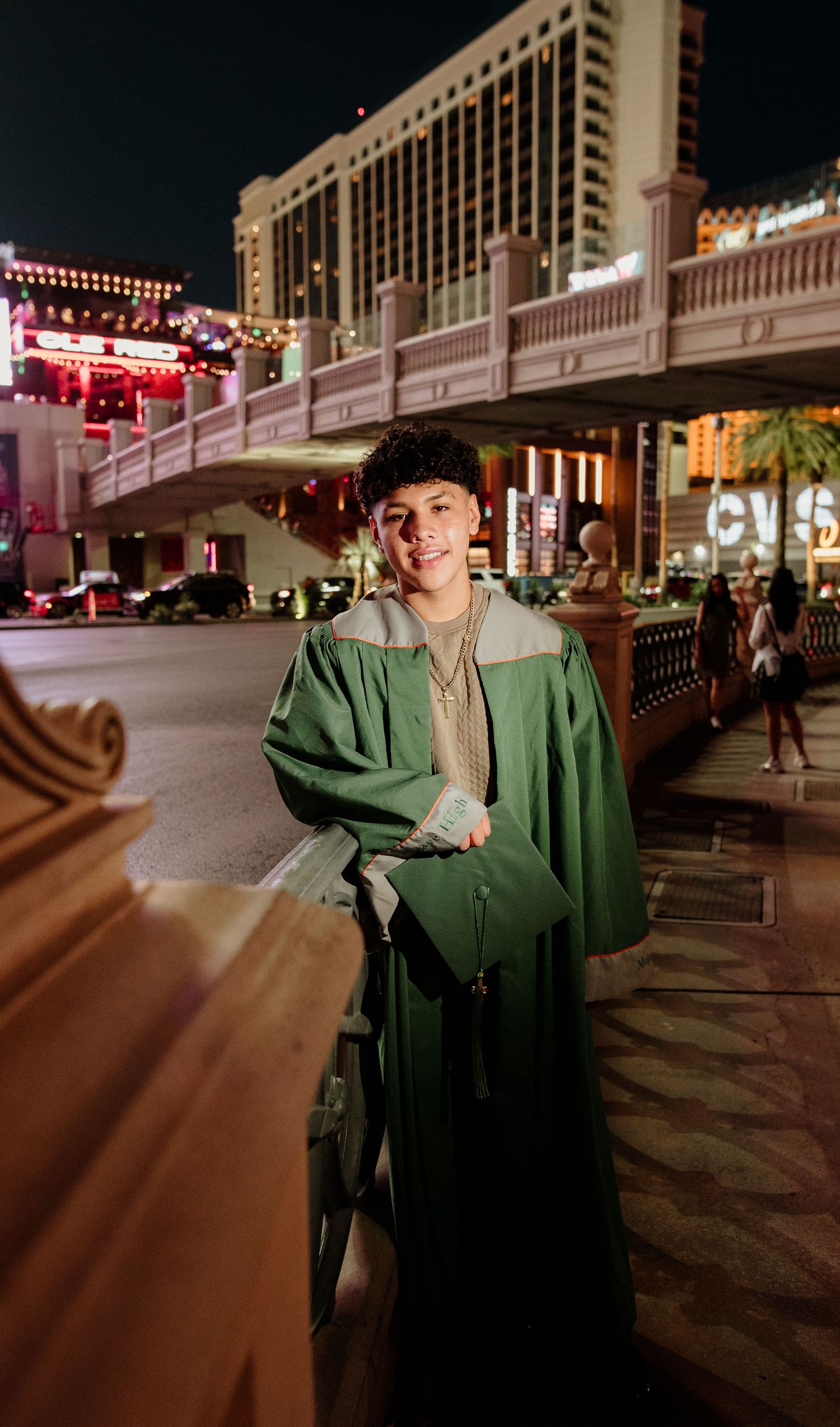 Young man in green graduation gown smiling on the Las Vegas Strip at night holding diploma.