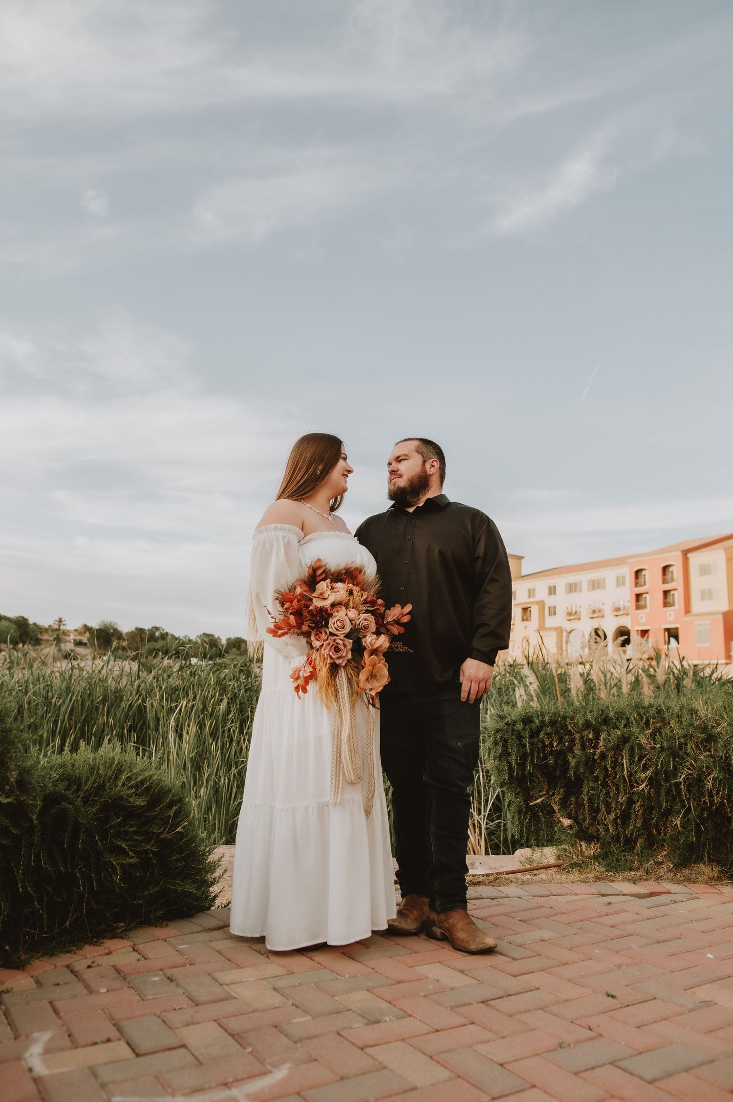 Bride and groom stand facing each other on brick path, bride holding autumn bouquet with tall grass behind.