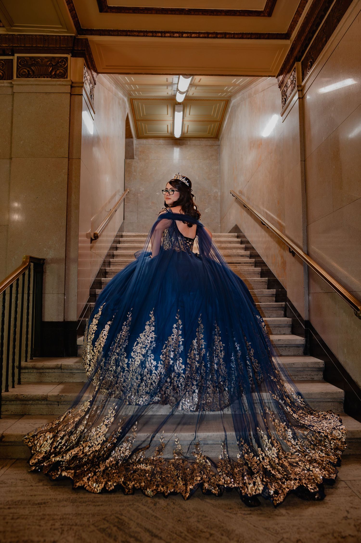 A young woman in a stunning navy blue and gold quinceañera ball gown poses on marble stairs in an elegant venue.