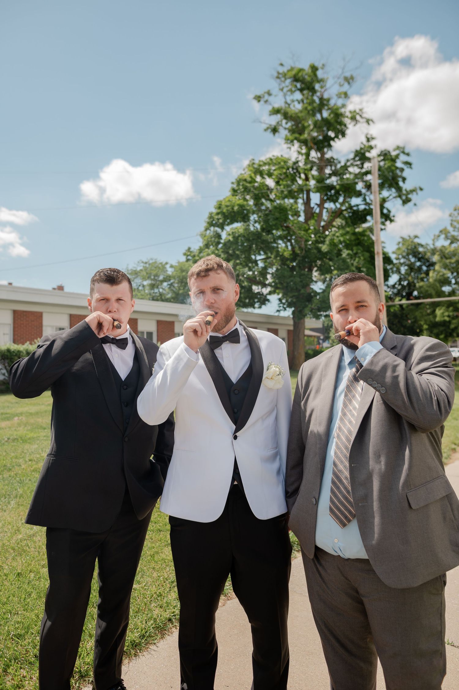 Three groomsmen in formal suits smoking cigars outdoors on a sunny wedding day.