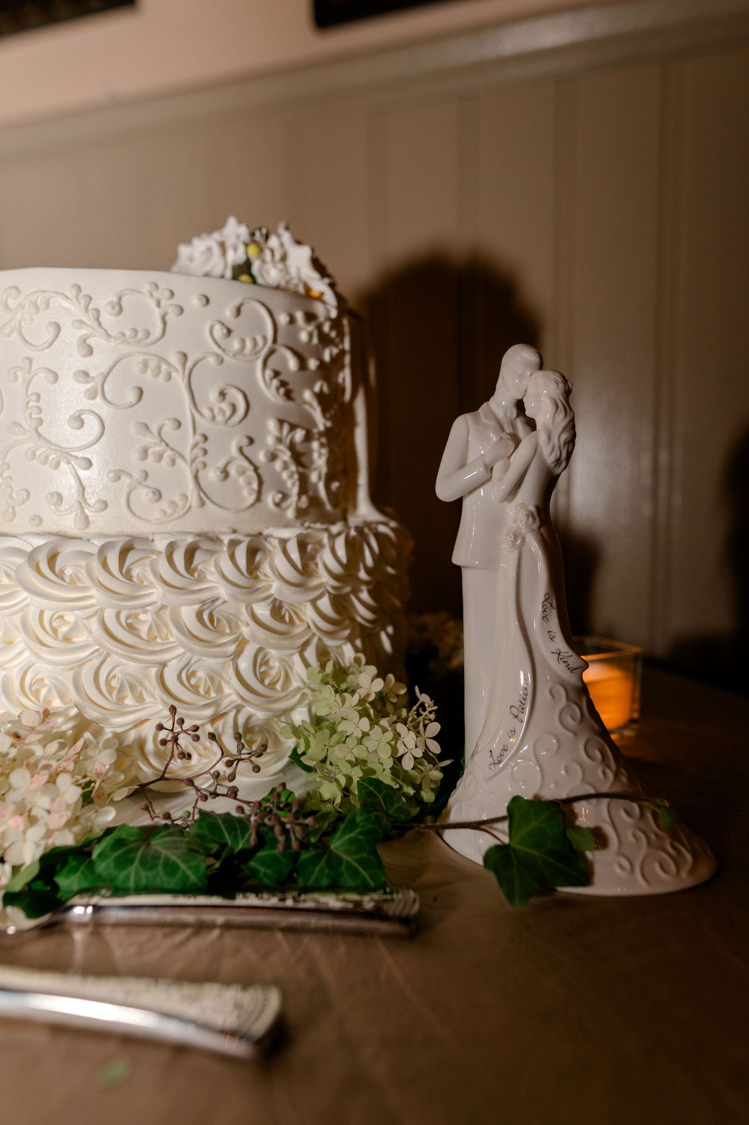 Elegant three-tier white wedding cake with intricate scroll designs beside a ceramic bride and groom cake topper.