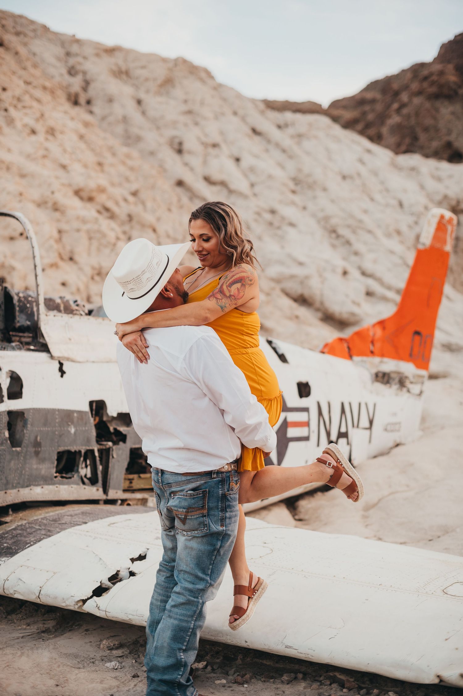 Man lifts woman joyfully near a vintage Navy airplane wreck in a desert canyon.