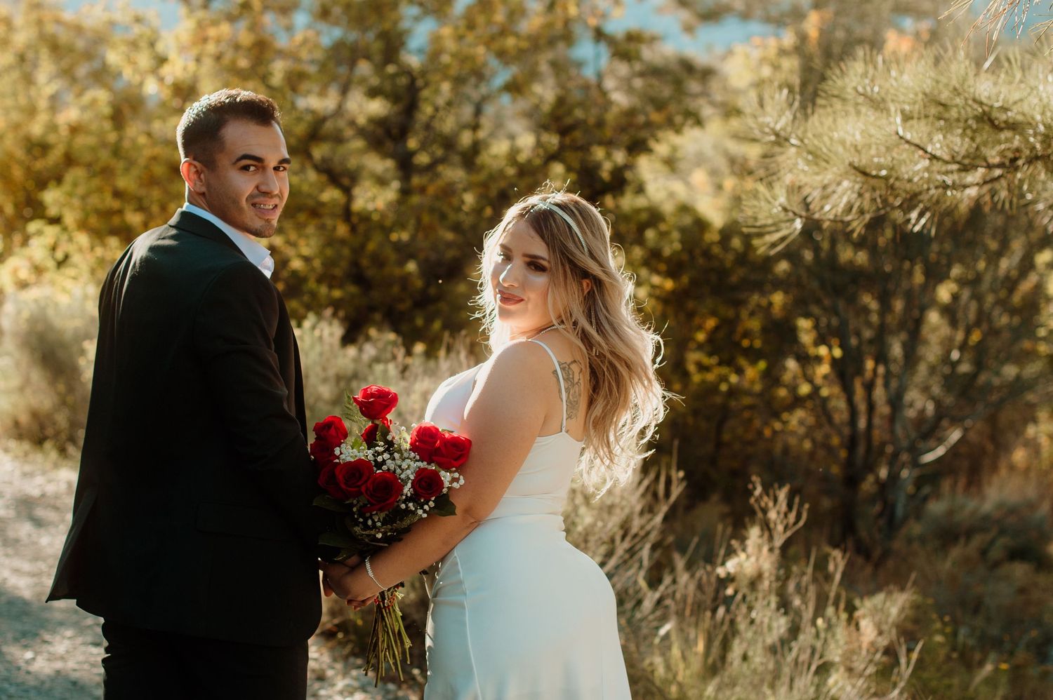 Bride in white dress holds red roses as groom in black suit smiles in desert setting.