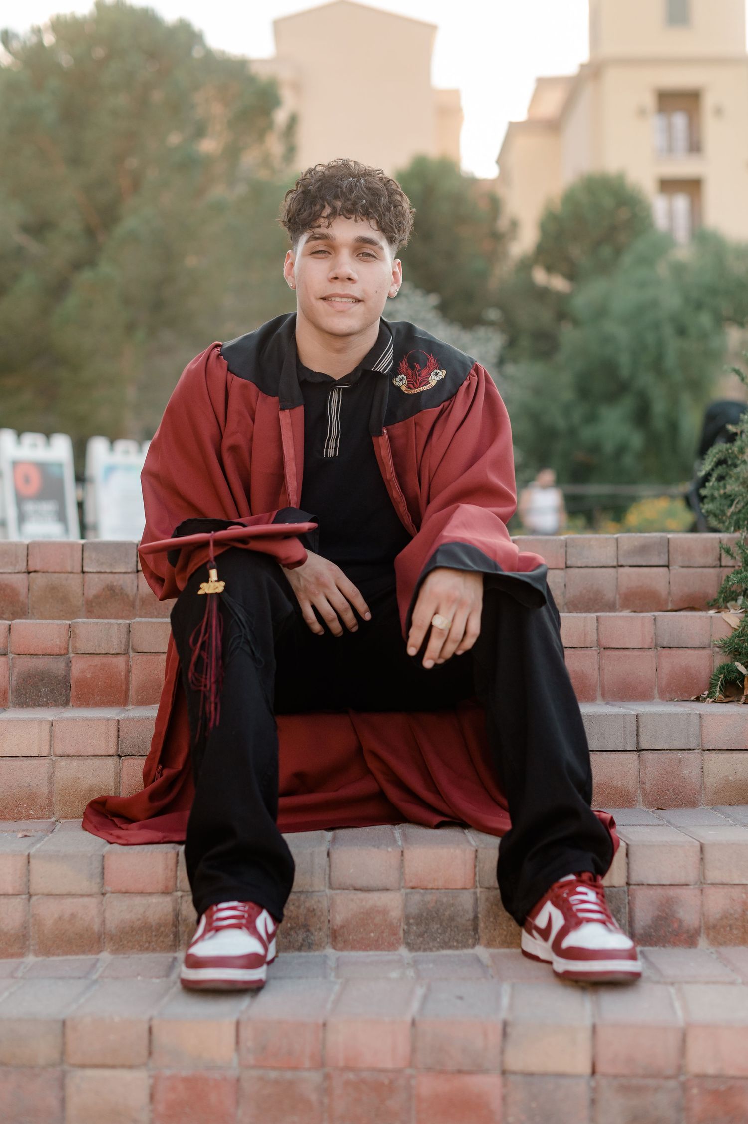 Graduate in red gown sits relaxed on brick steps holding cap with sneakers visible.