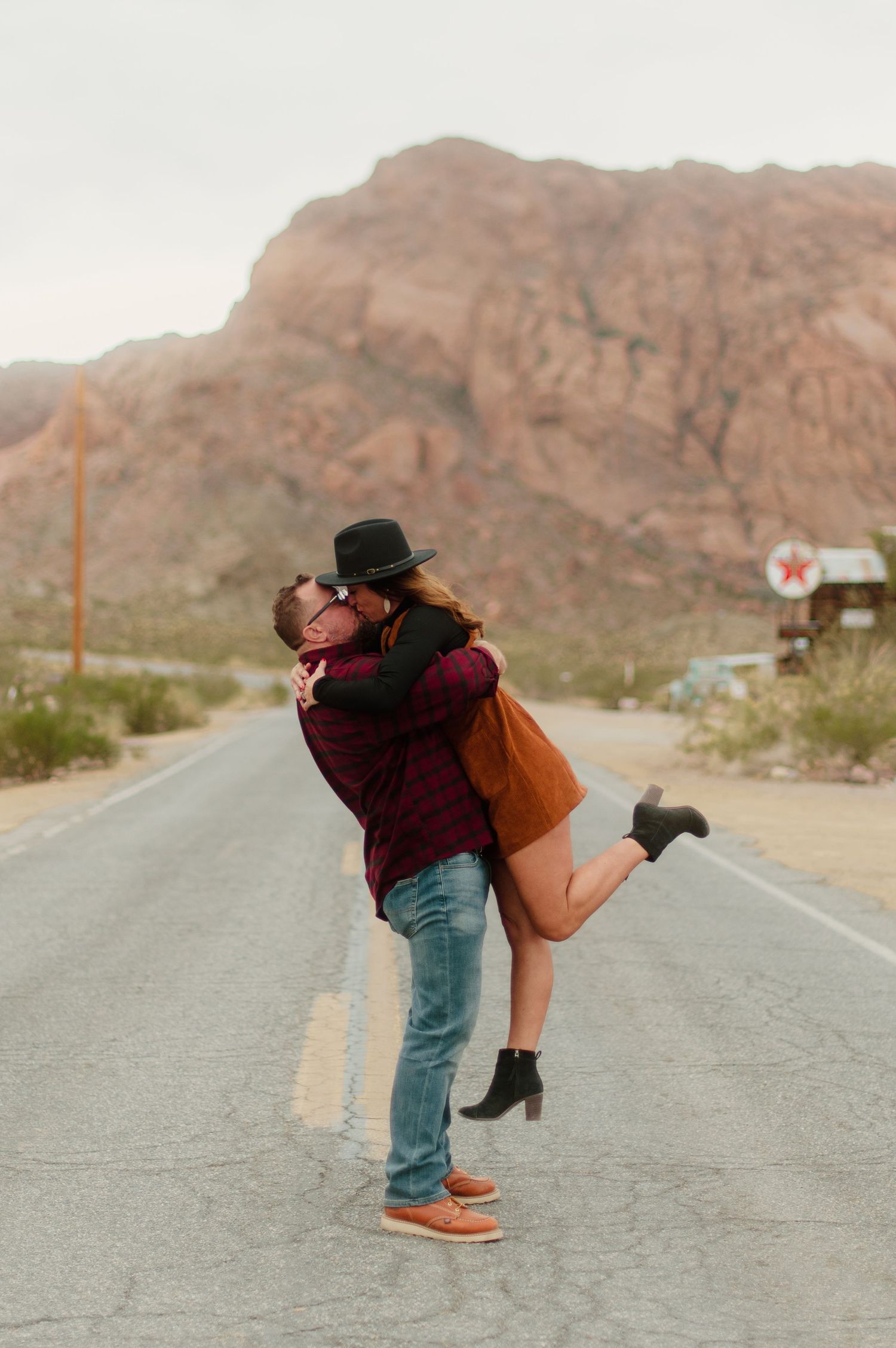 Man lifting woman in orange dress for a kiss on a scenic desert highway.