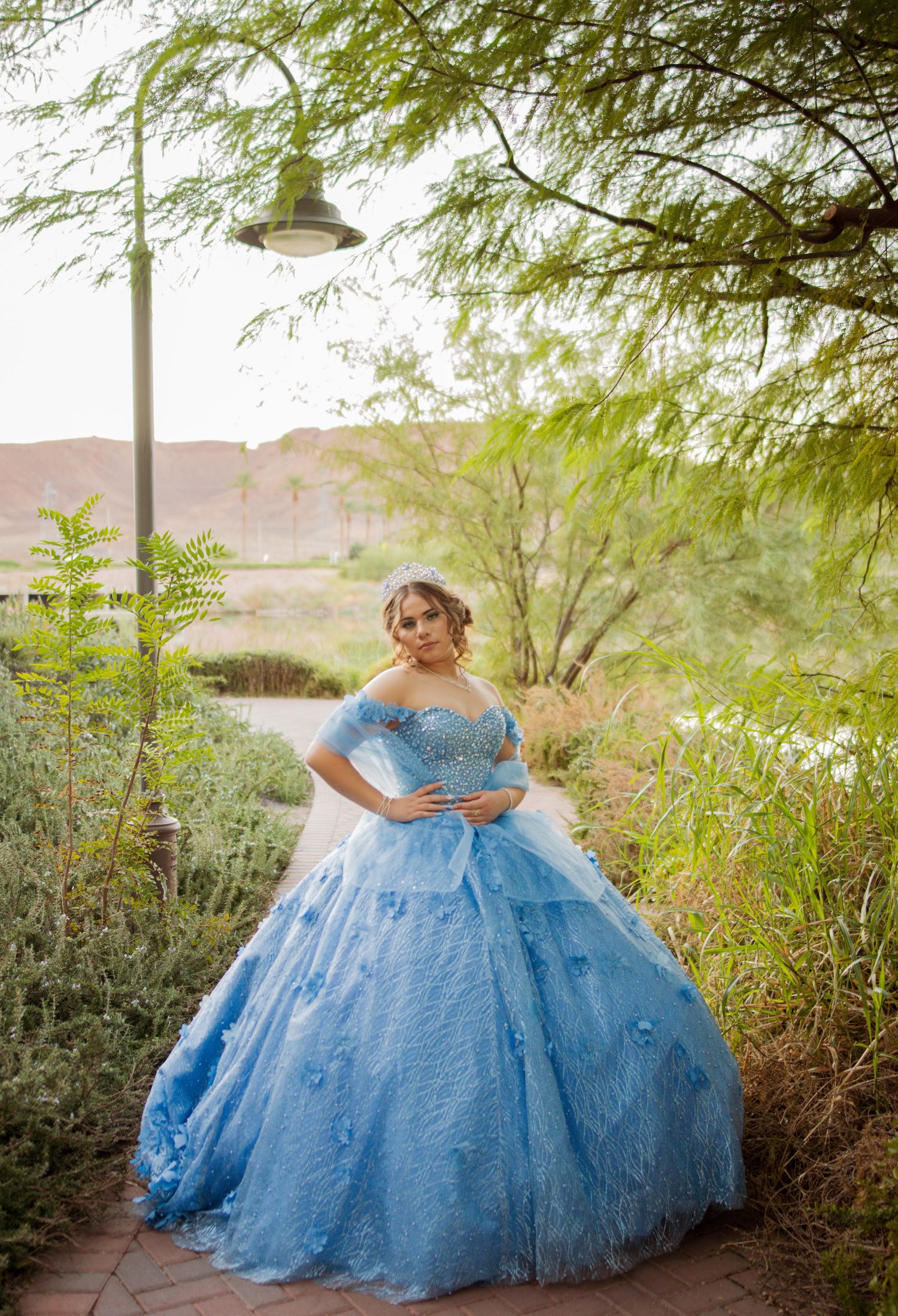 Young woman in a blue off-shoulder ball gown quinceañera dress poses in a lush garden pathway.