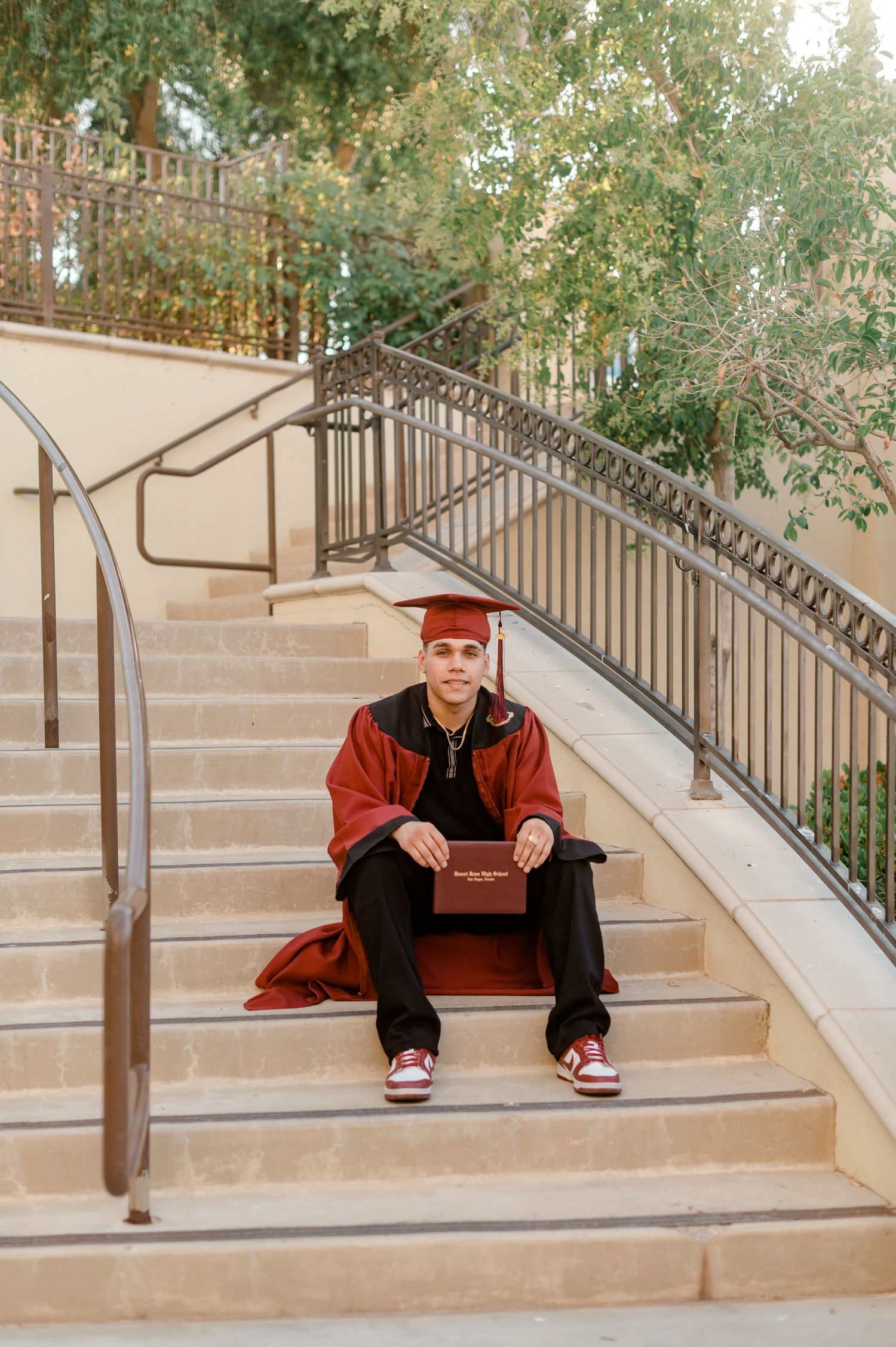 Graduate in red cap and gown sits on outdoor staircase holding diploma on campus.
