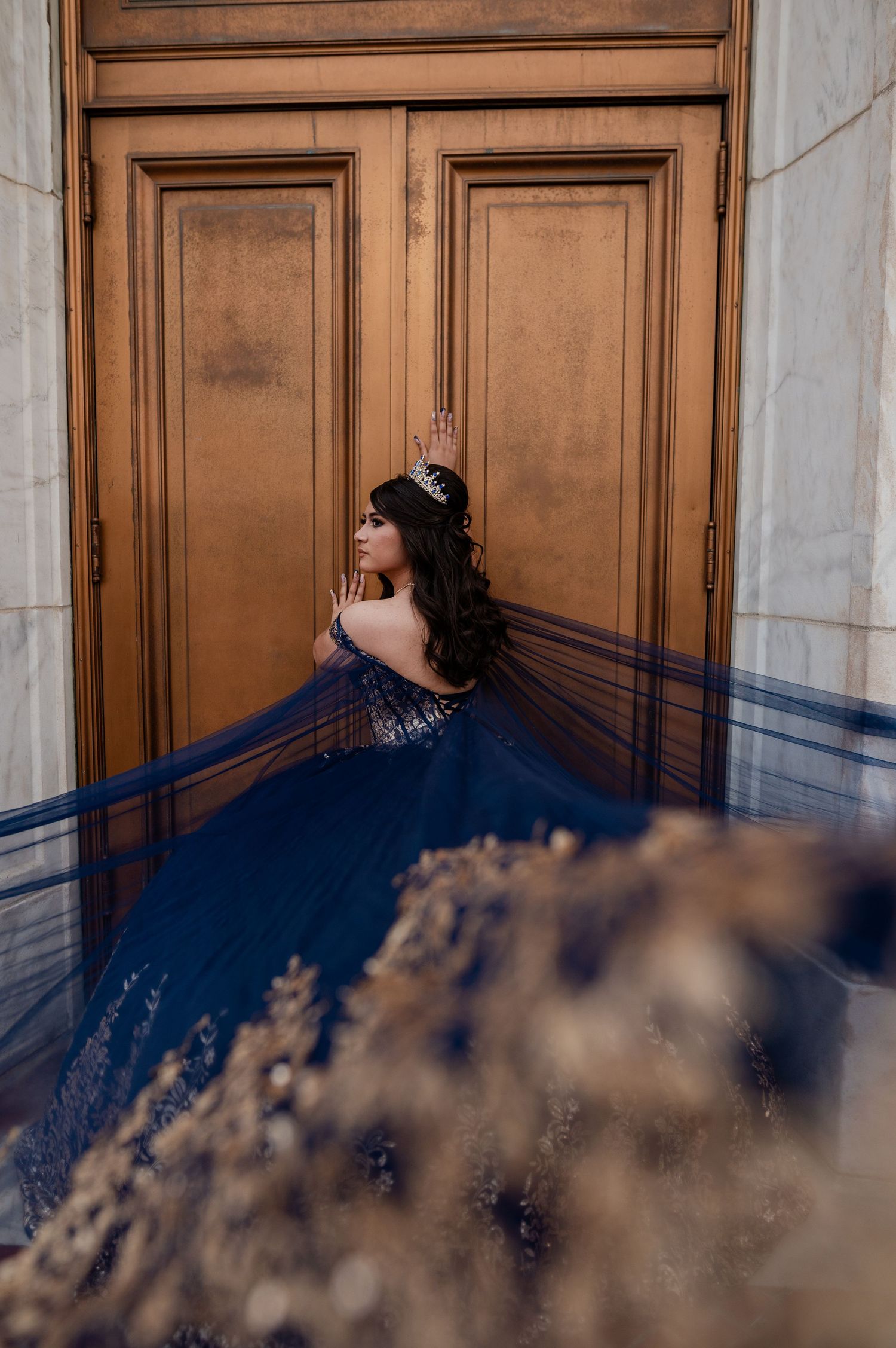 Young woman in navy blue quinceañera gown with tiara poses elegantly against ornate golden doors.