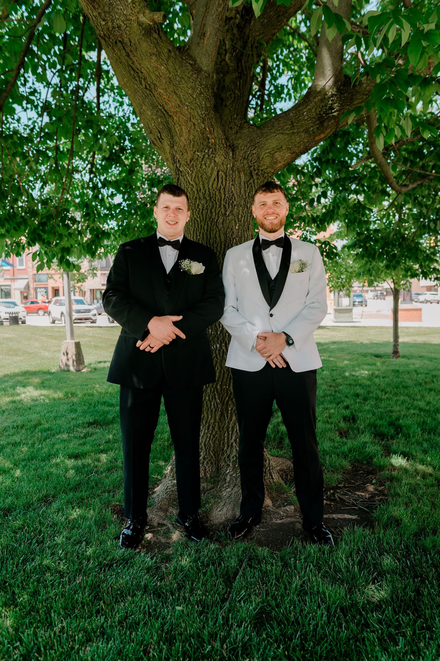 Two grooms in tuxedos pose together under a large tree on green grass at an outdoor wedding ceremony.
