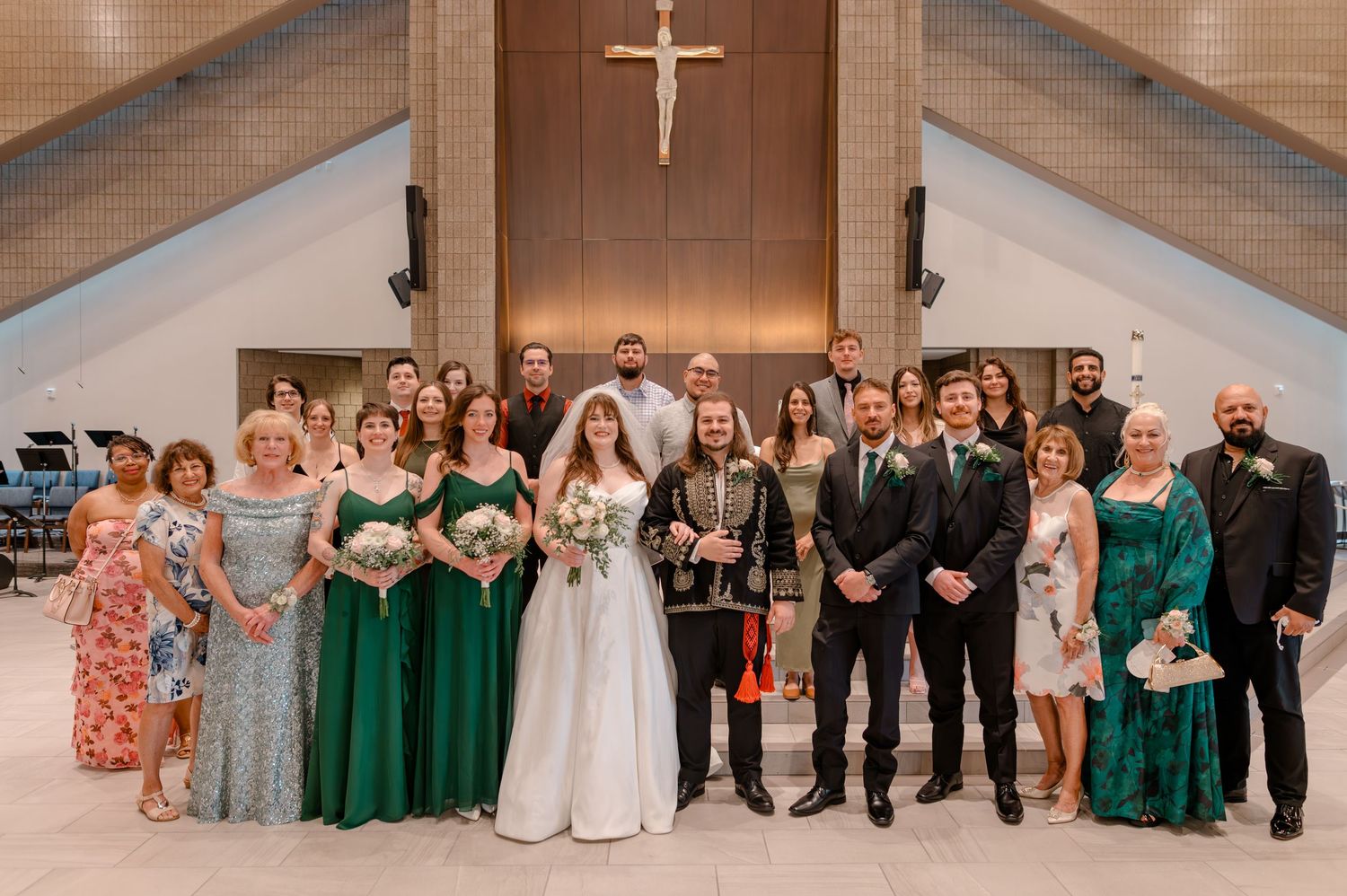 Wedding party in green dresses and suits posing together inside a church with a cross in the background.