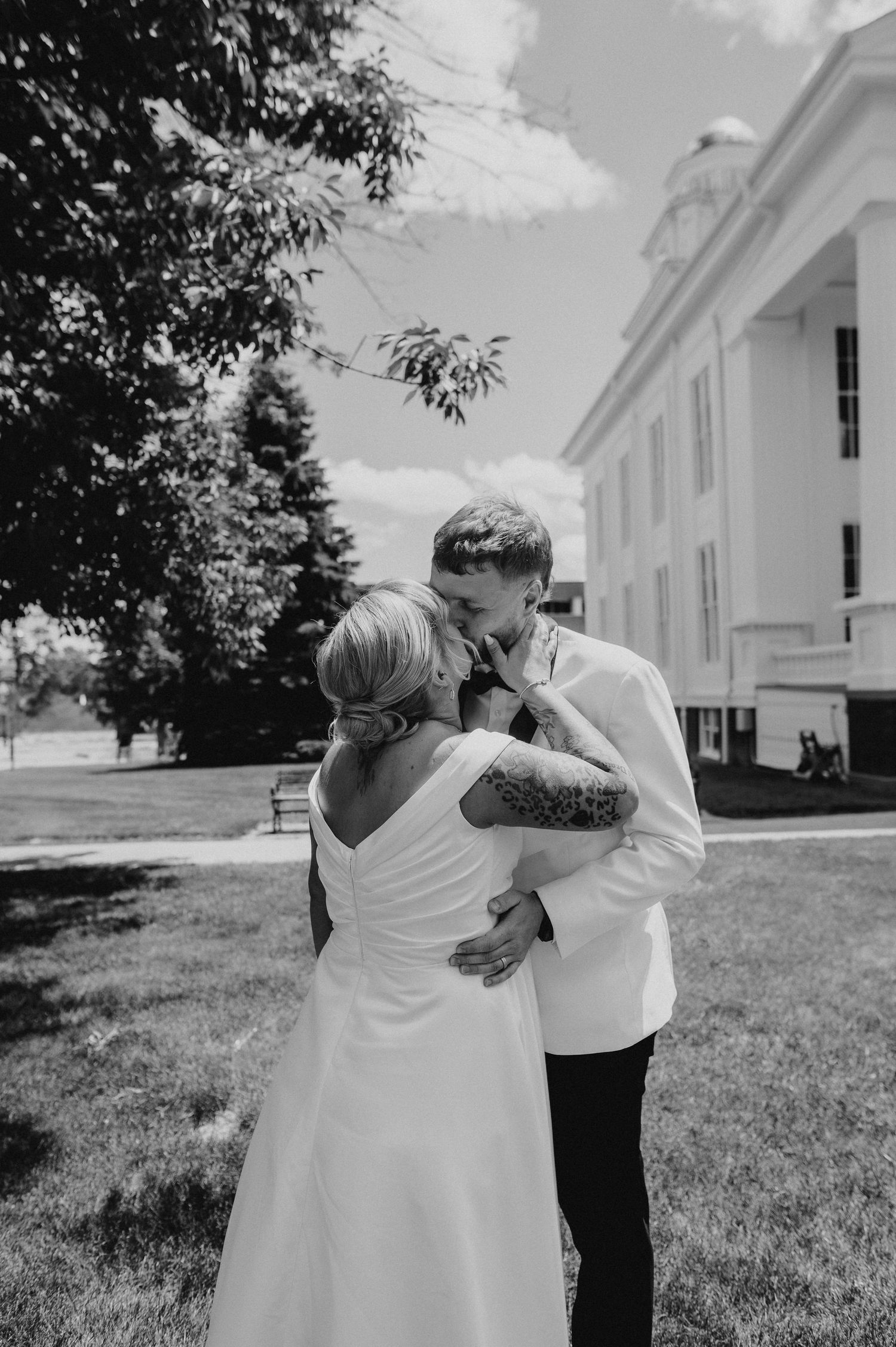 Black and white photo of tattooed bride and groom embracing outside a white colonial building.