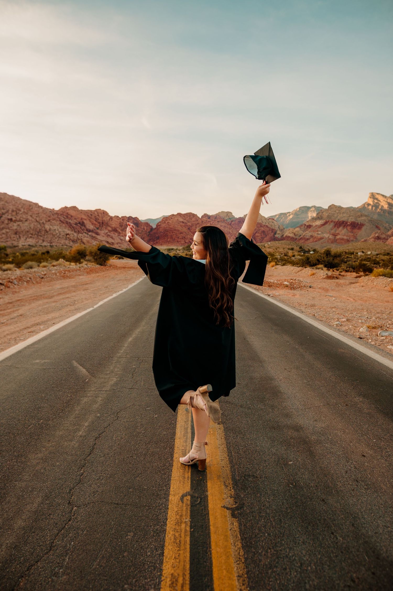 Graduate celebrates on desert road centerline, raising cap and arms toward Red Rock Canyon.