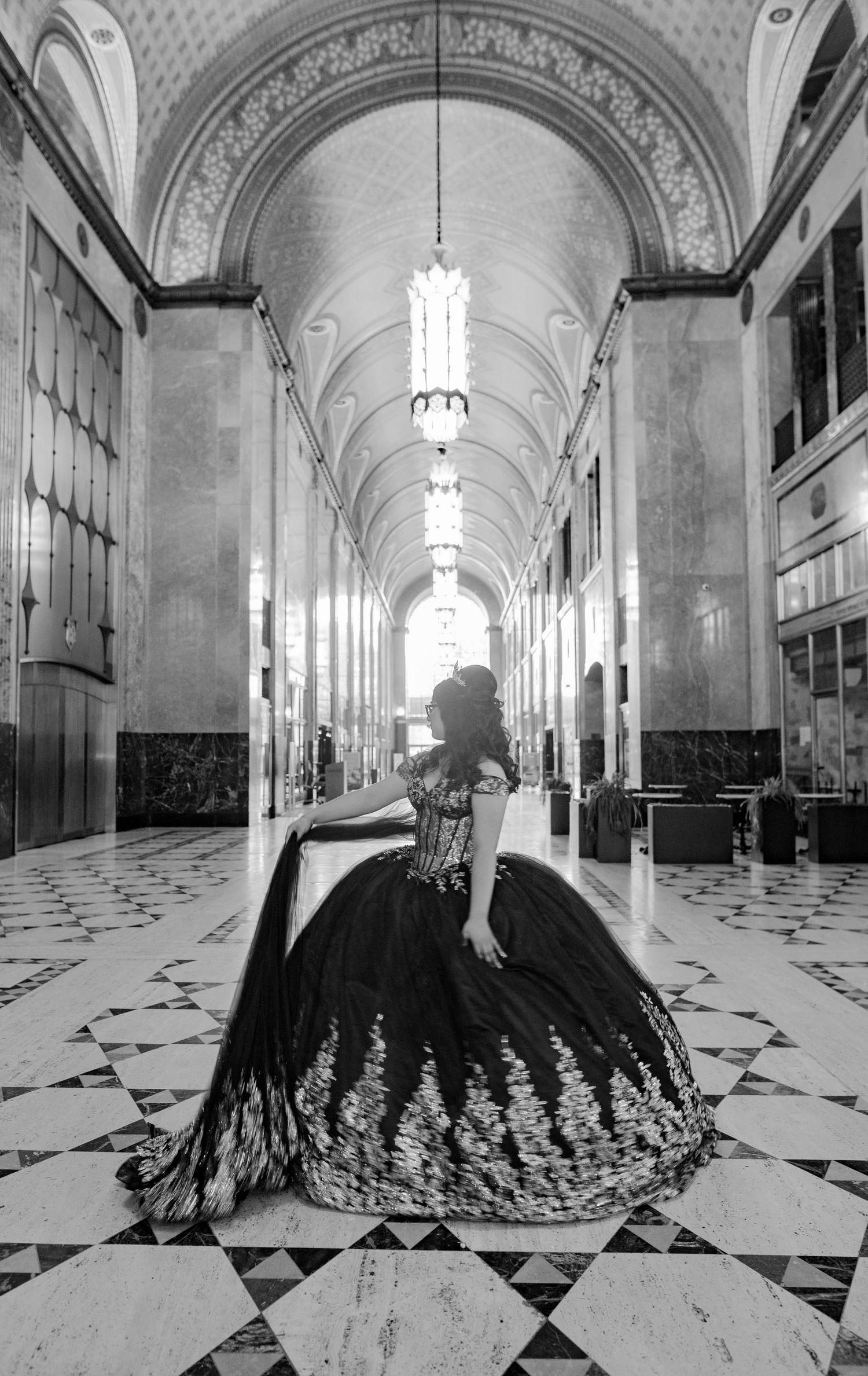 Black and white photo of a woman in an elegant ball gown posing in a grand ornate hallway with arched ceilings.