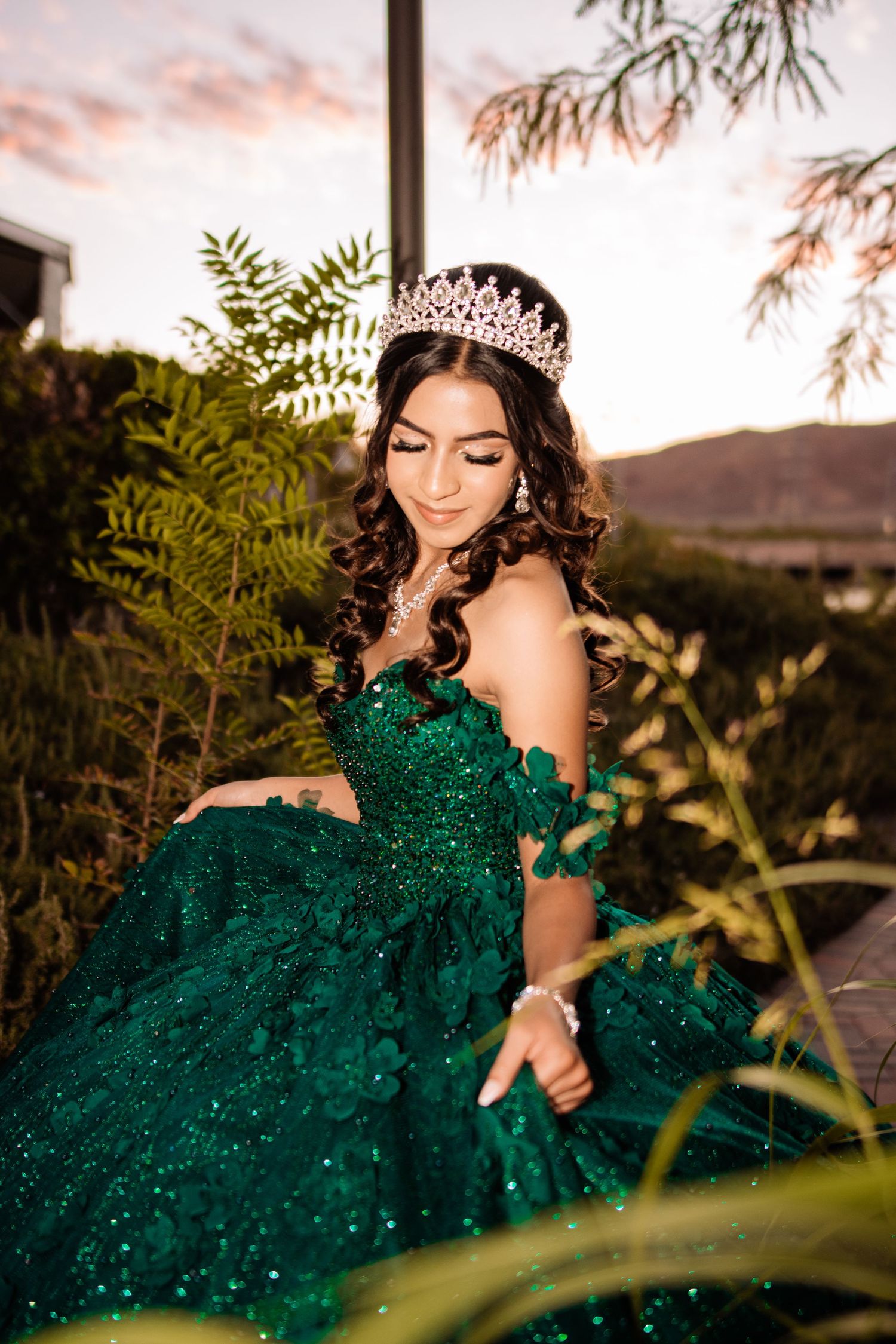 Young woman in emerald green sequined quinceañera gown with rose gold tiara poses among lush garden plants.